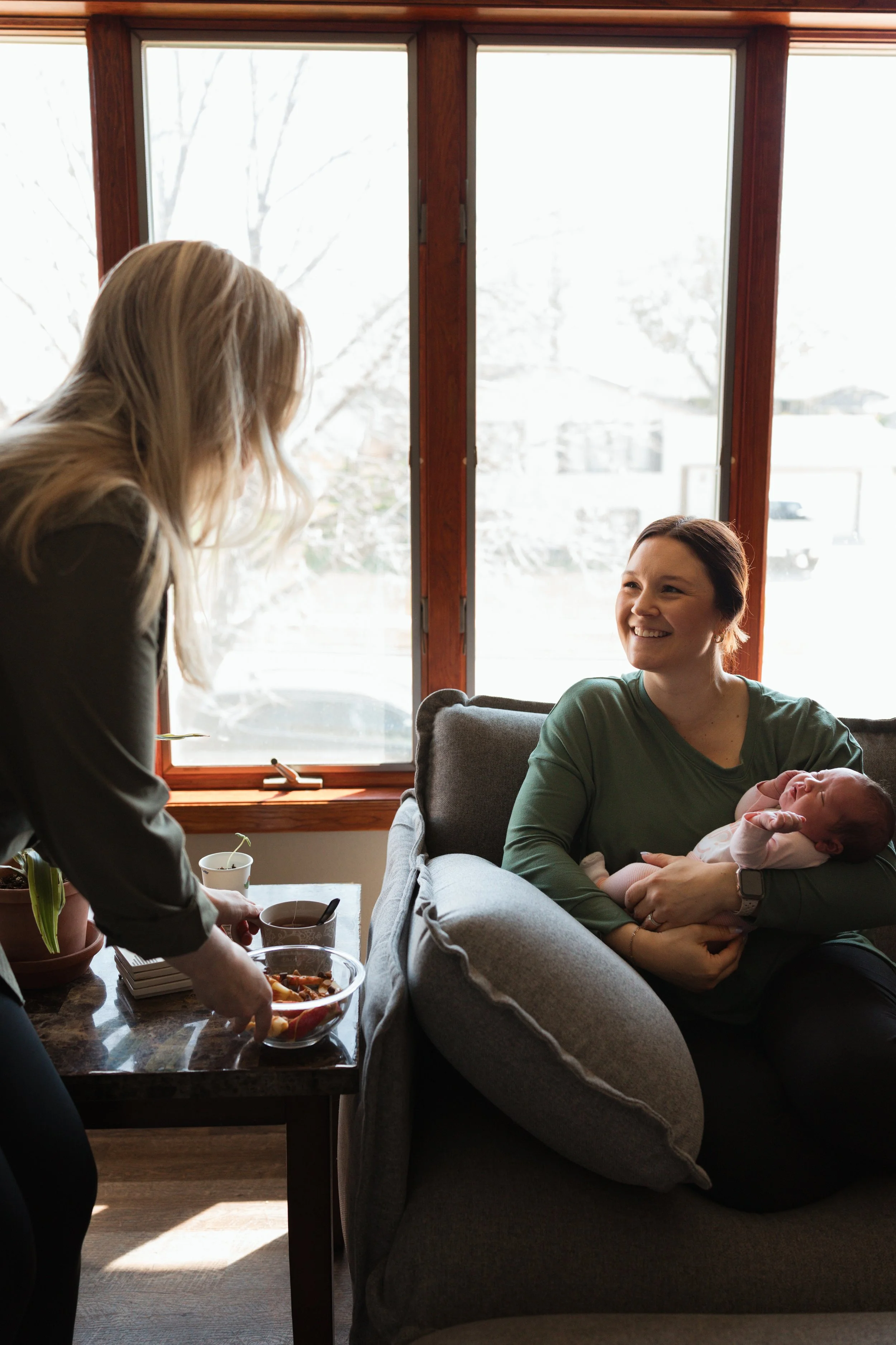 A woman sitting on a gray couch holding a newborn baby while another woman stands nearby with a plate of food and drinks on a table. They are in a room with large windows overlooking a snowy landscape outside.