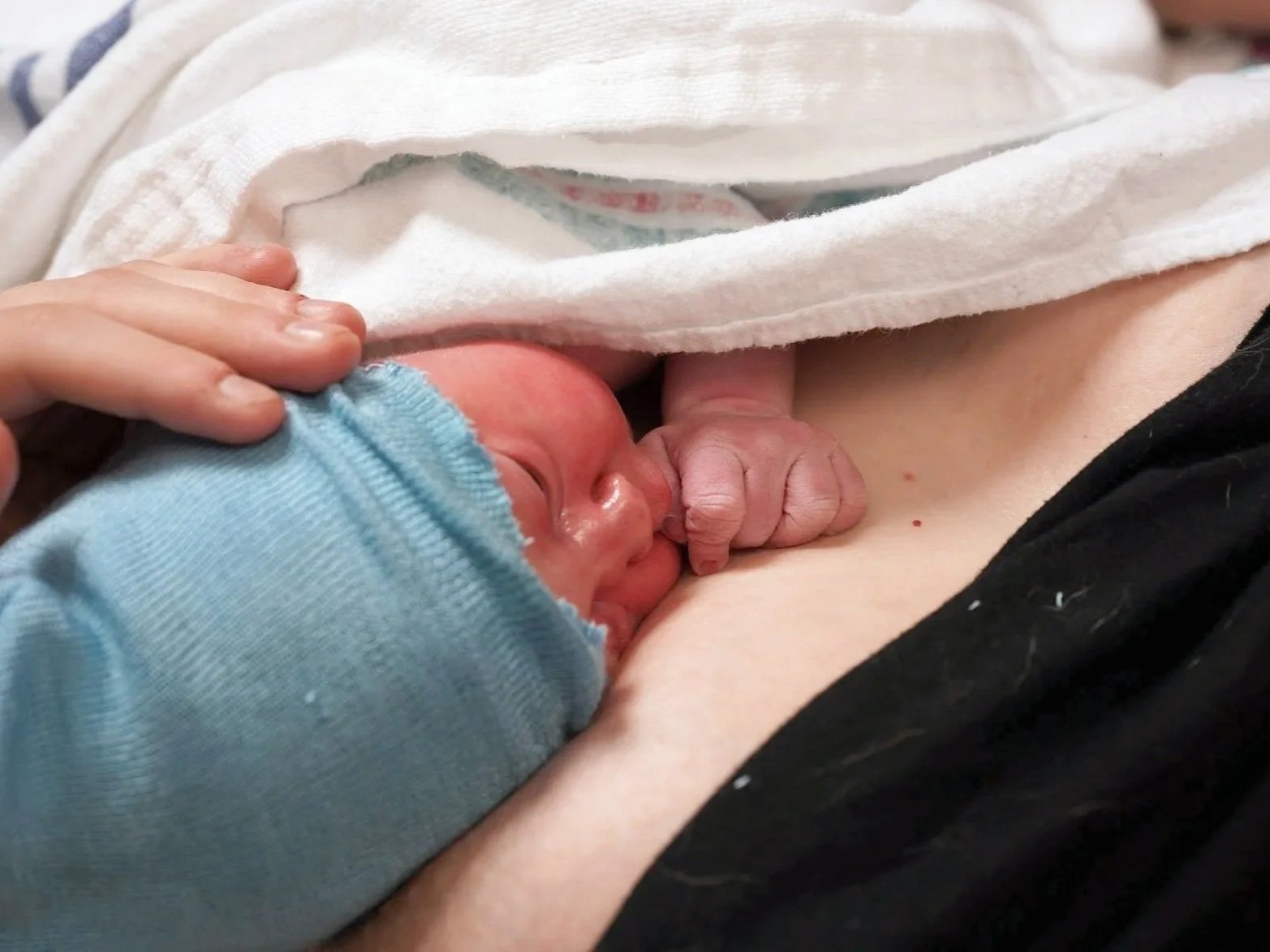 A newborn baby nursing at a mother's breast, covered partially by a white cloth, with the baby's face and hand visible, wearing a blue outfit.