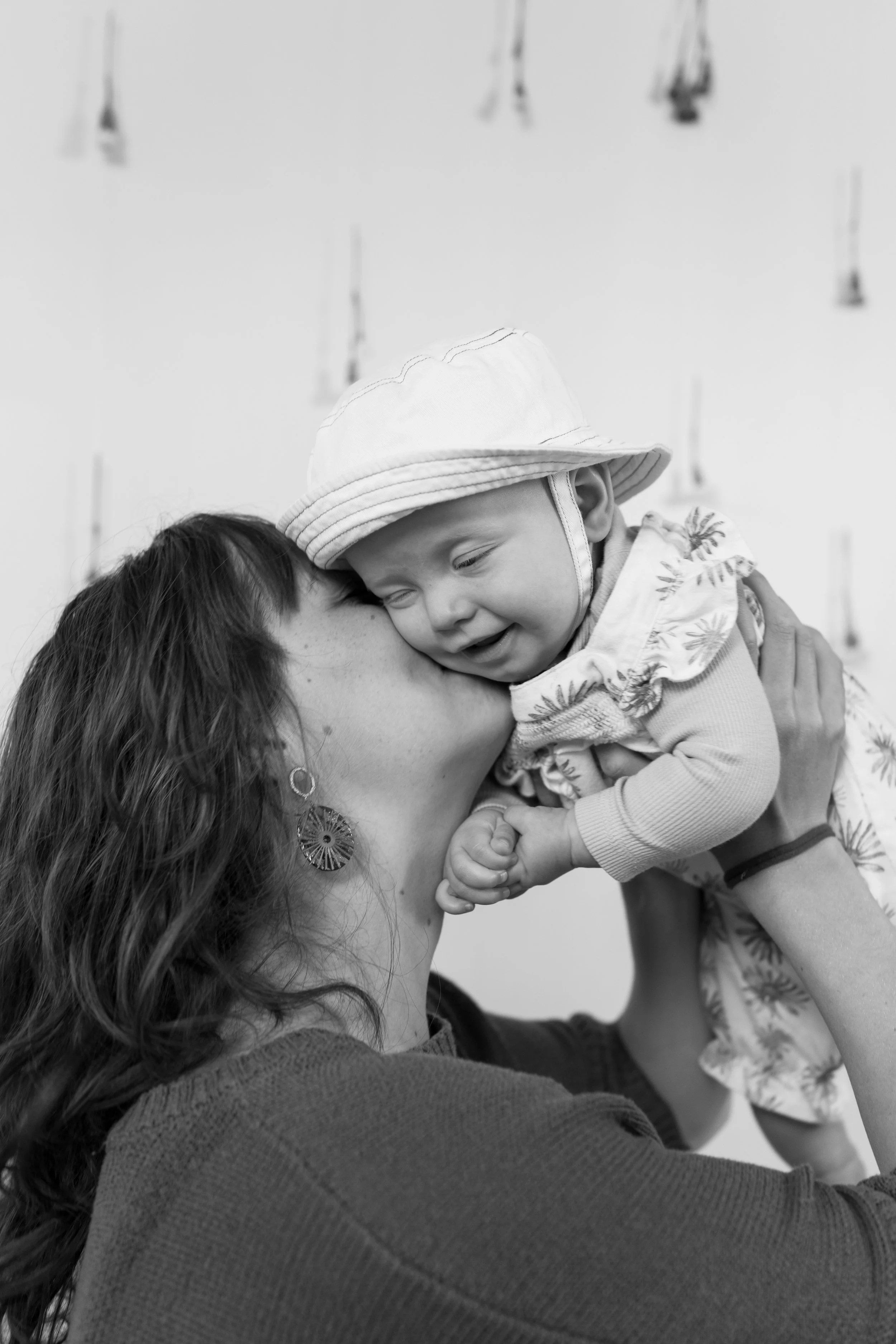A woman with long wavy hair, wearing large earrings and a sweater, holding a smiling young child with a hat and floral shirt, in a close embrace.