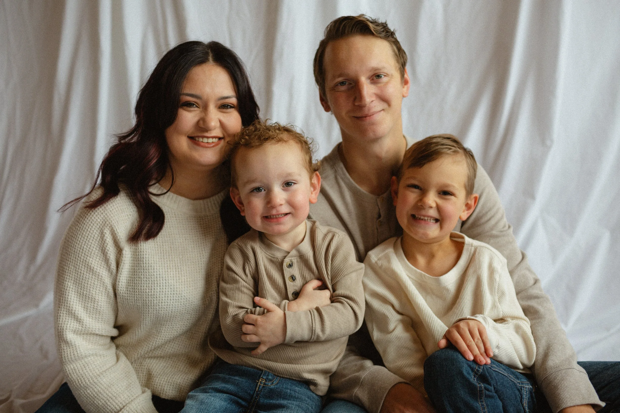 A family of five, including a woman, two young boys, and two teenage boys, sitting together on a sofa, smiling for the camera against a white curtain backdrop.
