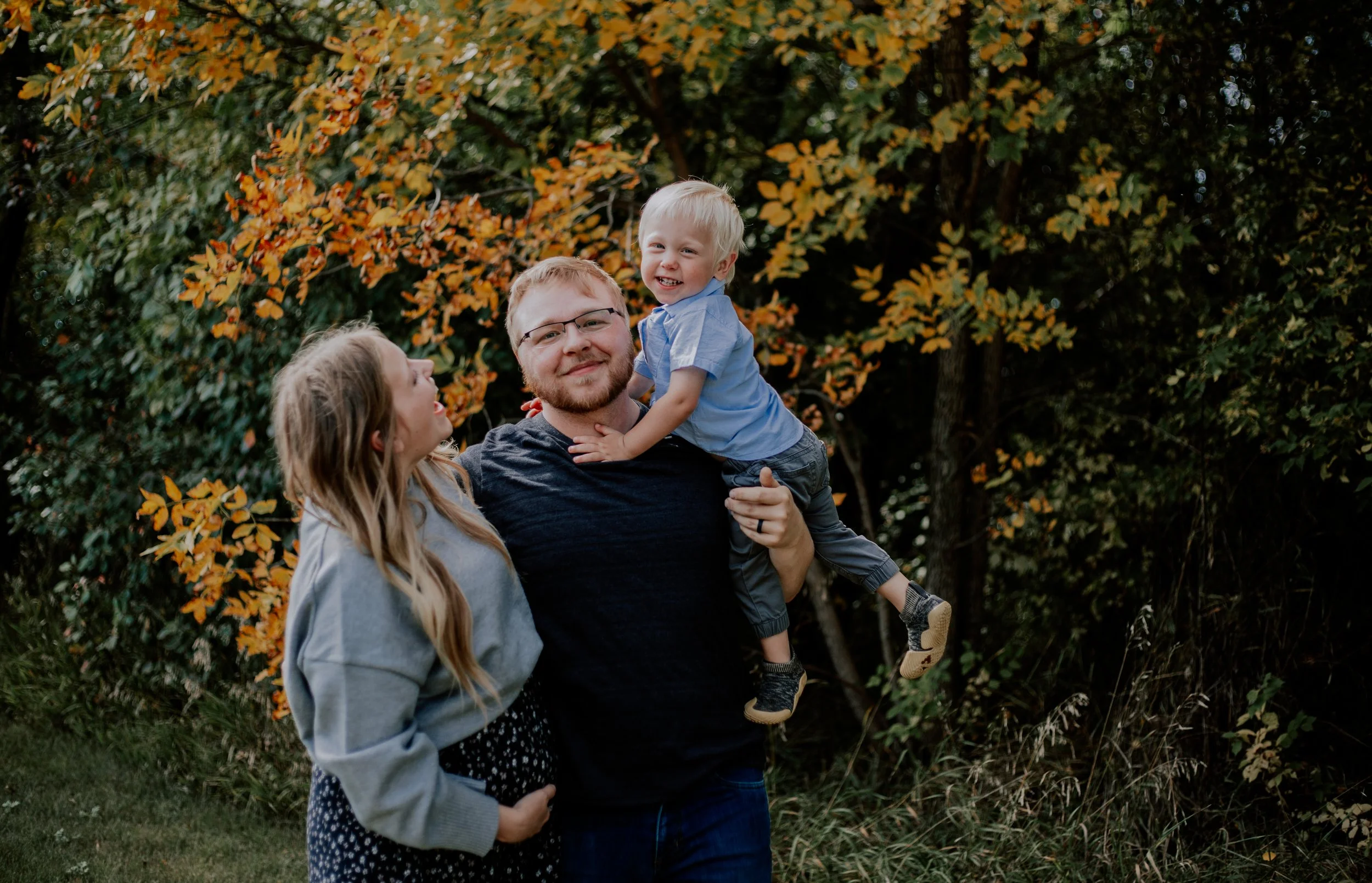 A man holding a young boy in his arms, standing outdoors with trees and autumn leaves in the background. A girl stands nearby, looking at the boy and smiling.