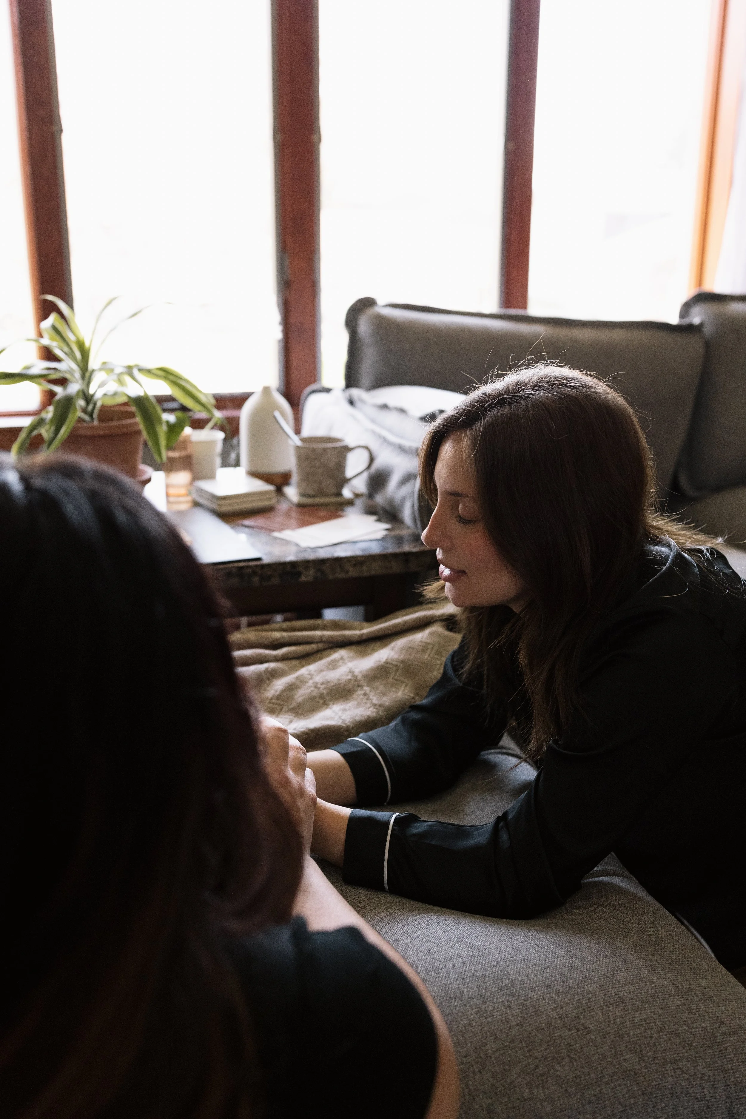 A woman with brown hair is sitting on a couch, gently holding a child's arm, with her eyes closed, in a cozy living room with a table, potted plant, and window in the background.