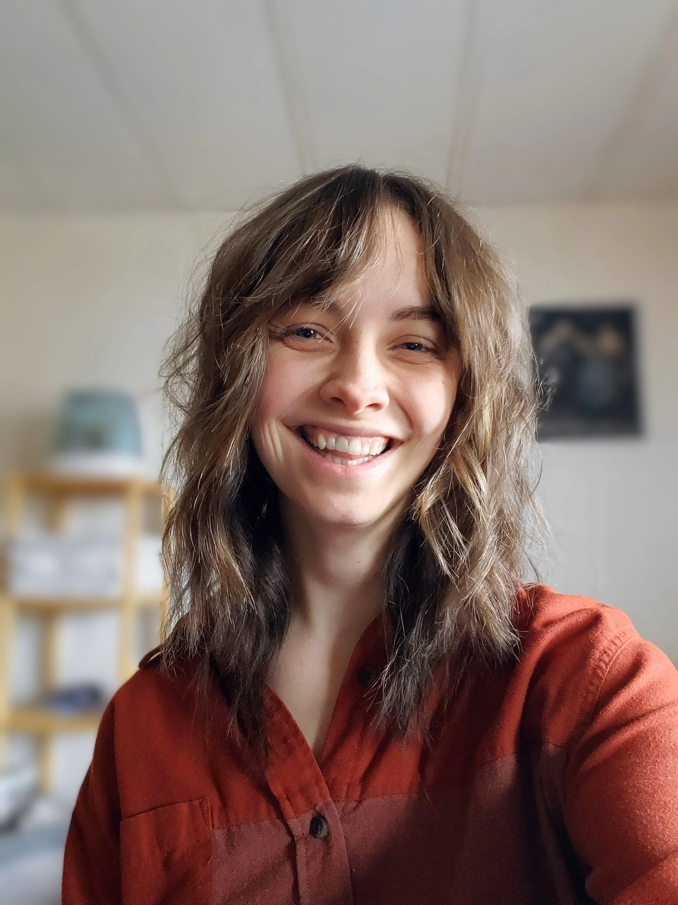 A smiling woman with wavy, shoulder-length hair and a reddish-brown shirt. The background shows a blurred room with a shelf and a framed picture on the wall.