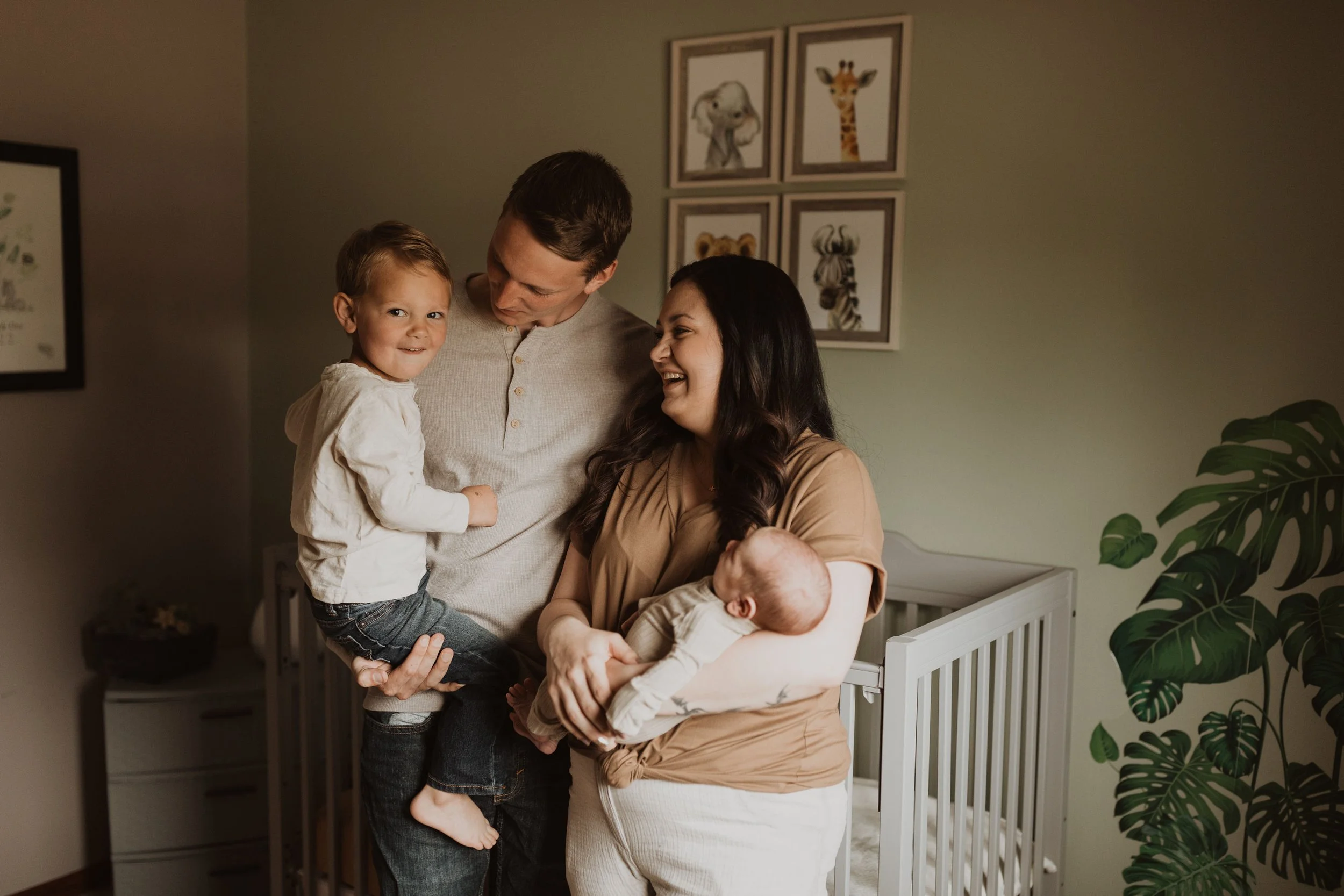 A family in a nursery room, including two adults and two children, with framed animal prints on the wall and a large green houseplant.