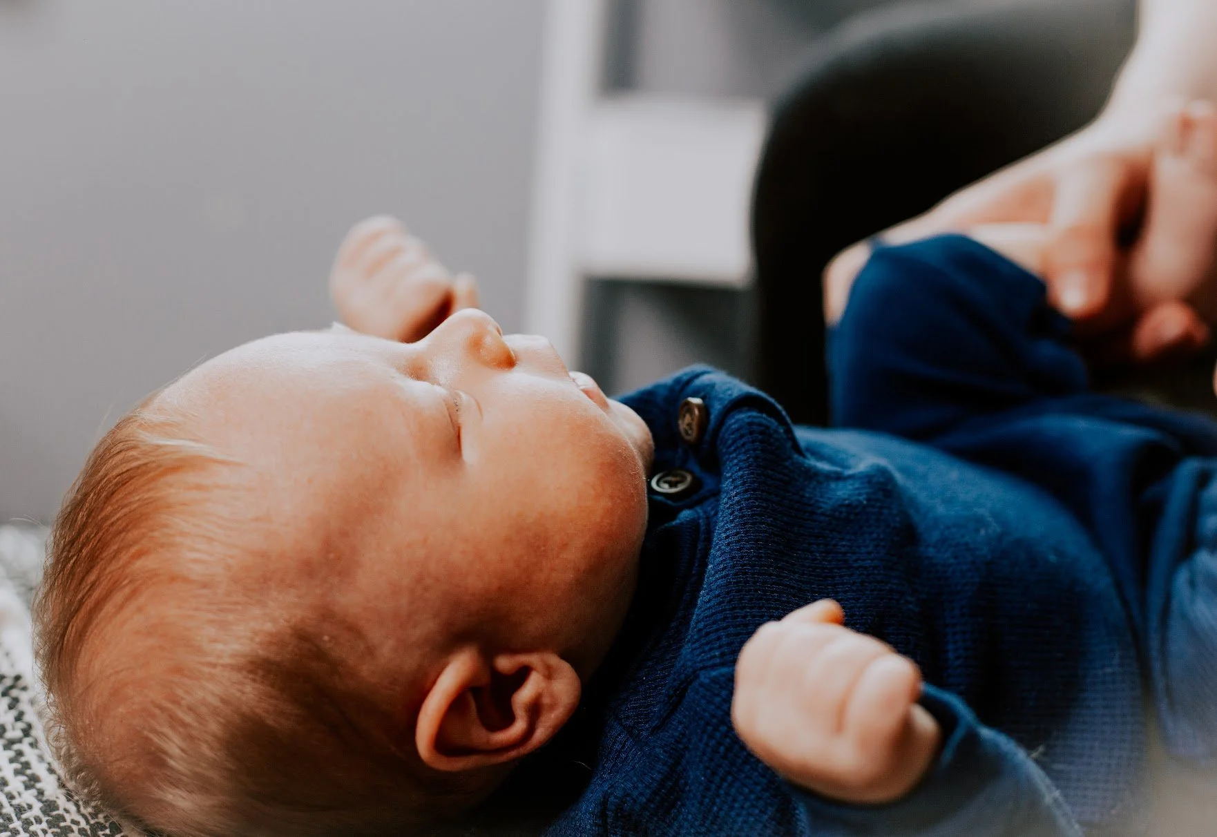 Close-up of a sleeping baby with light skin and red hair, lying on a gray and white blanket, wearing a dark blue shirt.