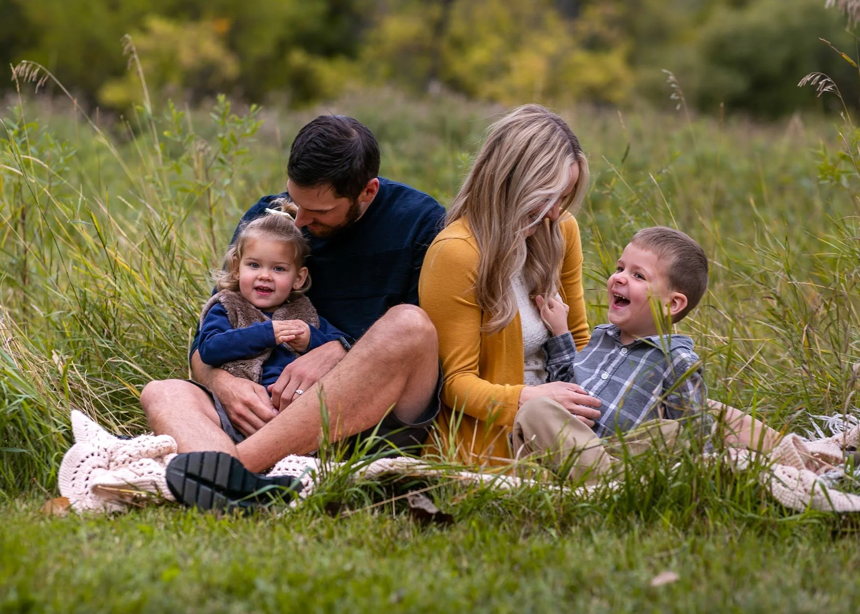 A family of four sitting in tall grass outdoors, smiling and laughing. The father has a young girl on his lap, and the mother is sitting next to a young boy, both children are looking happy and engaged.