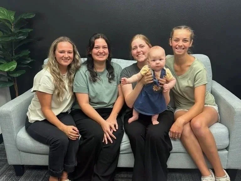 Five women sitting on a light gray sofa, smiling at the camera. One woman is holding a baby. There is a large green plant on the left side and a dark wall behind them.