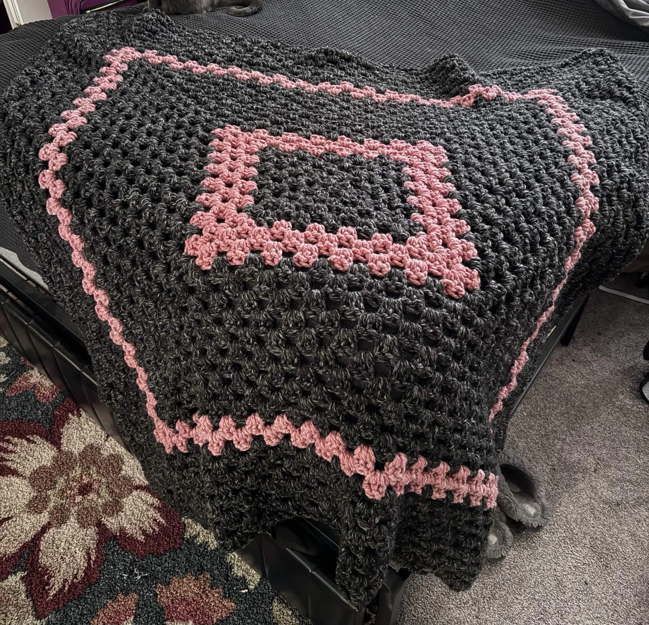 A black crocheted blanket in charcoal and pink, draped over a bed with a black bedspread.