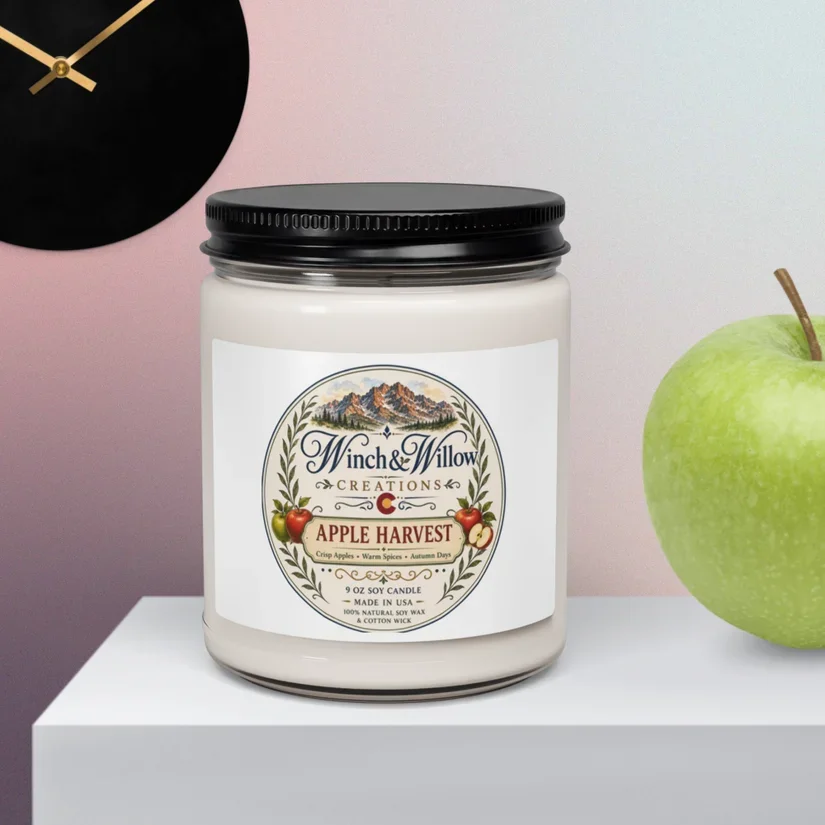 A white candle in a jar with a black lid, labeled Winch & Willow Creations Apple Harvest. The label features a mountain landscape and apples, and mentions it is made of natural soy wax with a cotton wick. The candle is on a white platform next to a green apple and a black clock on a pink wall.