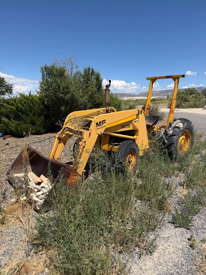 A yellow tractor abandoned in a field, overgrown with weeds and plants, with a clear blue sky and some distant hills in the background.