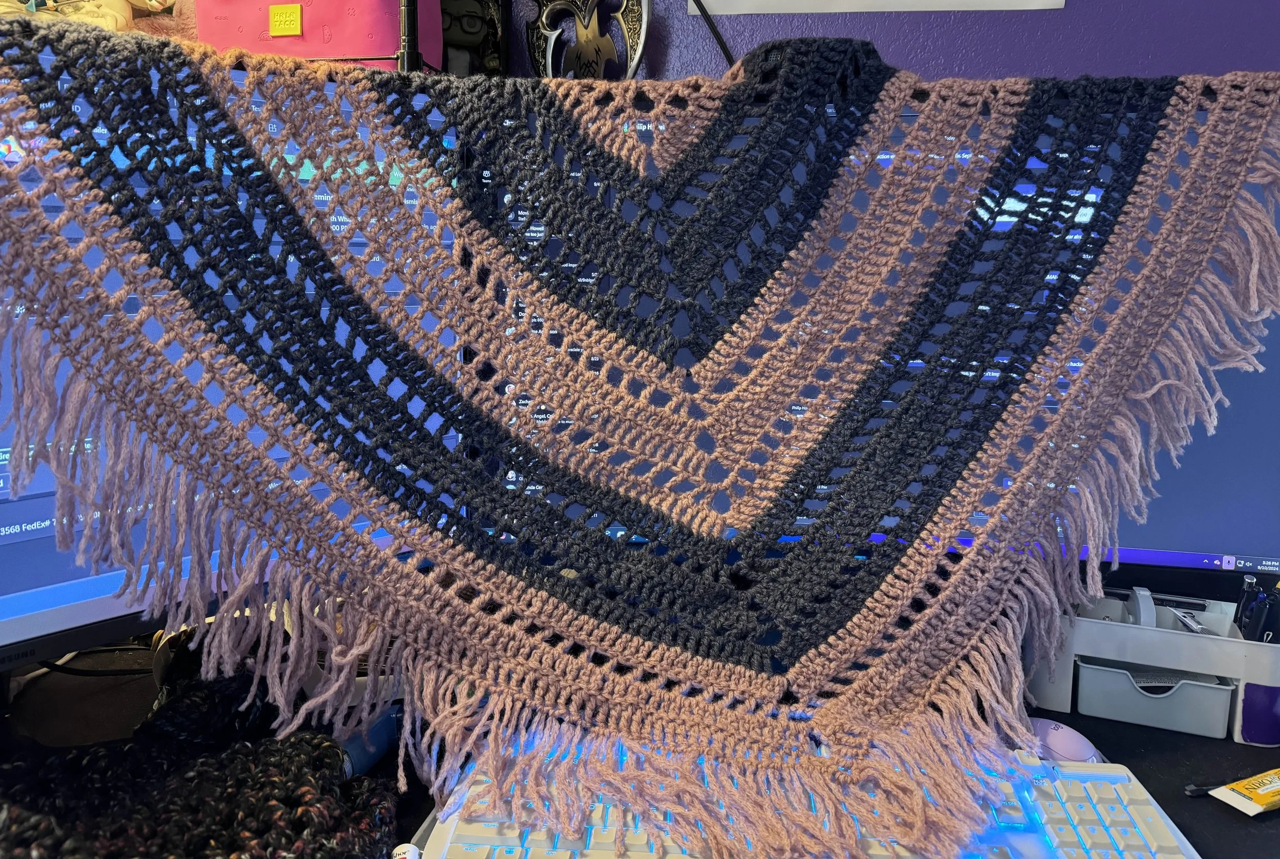 A large, open crochet shawl with a geometric pattern in pink and blue draped over a computer monitor.
