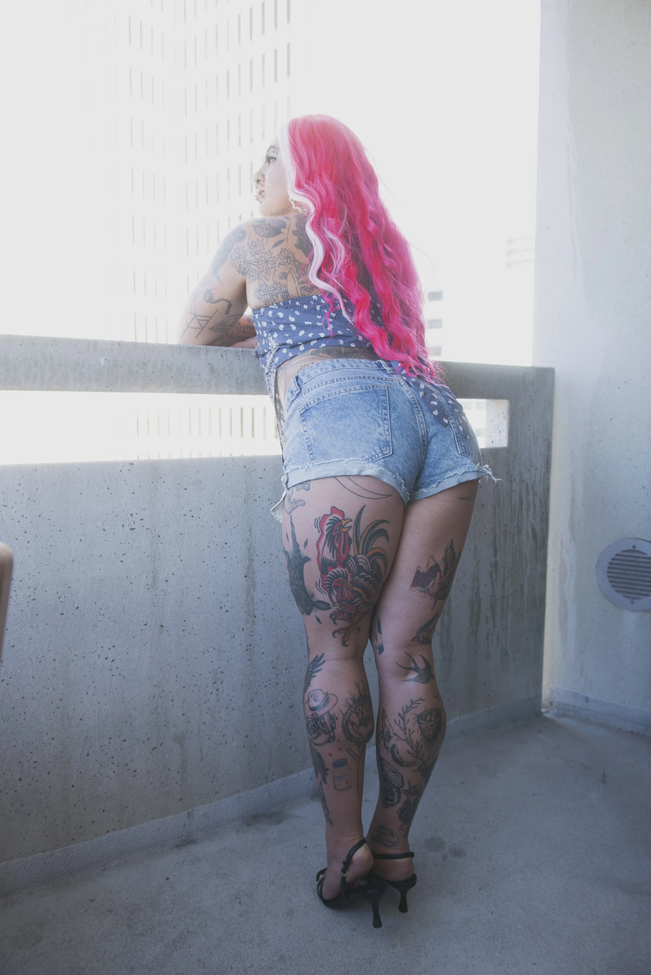 A ftm escort with pink hair leaning on a concrete balcony railing, wearing a blue patterned top, denim shorts, and black high-heeled shoes, with tattoos covering her arms and legs, on a balcony of a building. Bay area escort for hire 
