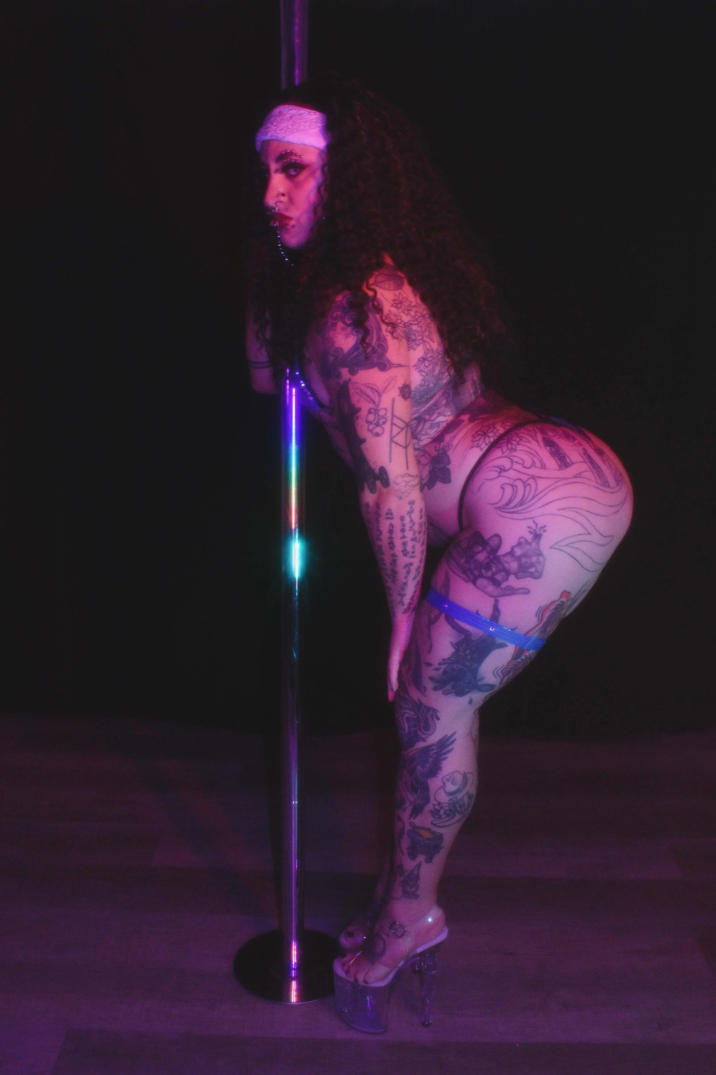 A trans stripper with tattoos wearing clear platform heels and high-heeled sandals, leaning on a pole in a dimly lit environment with purple neon lighting.