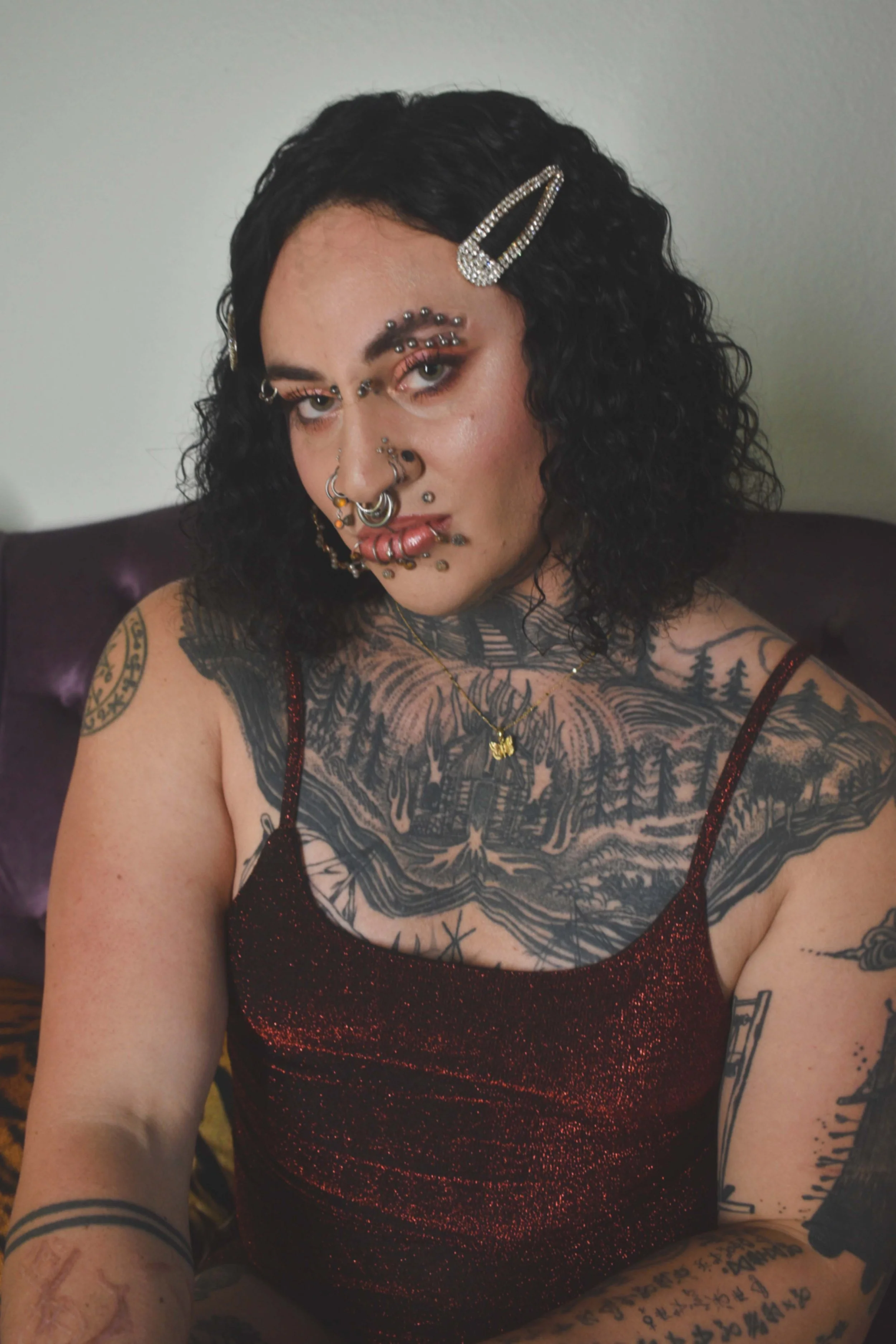 dark, short, curly hair decorated with rhinestone hair clips. They have multiple facial piercings, including eyebrows, nose, and lips, and are wearing makeup  They have tattoos on their chest, arms, and shoulders, and are wearing a red, sparkly spagh