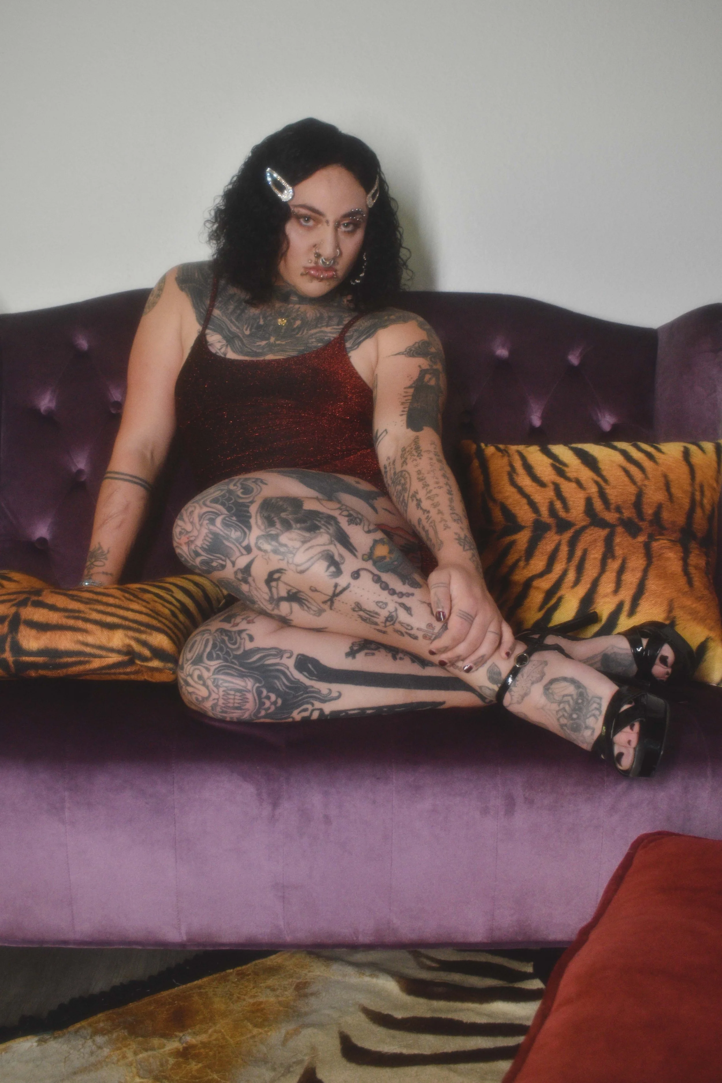 A trans stripper with black curly hair, multiple tattoos, and facial piercings sitting on a purple tufted sofa, wearing a red dress and black sandals with a tiger-striped cushion nearby.