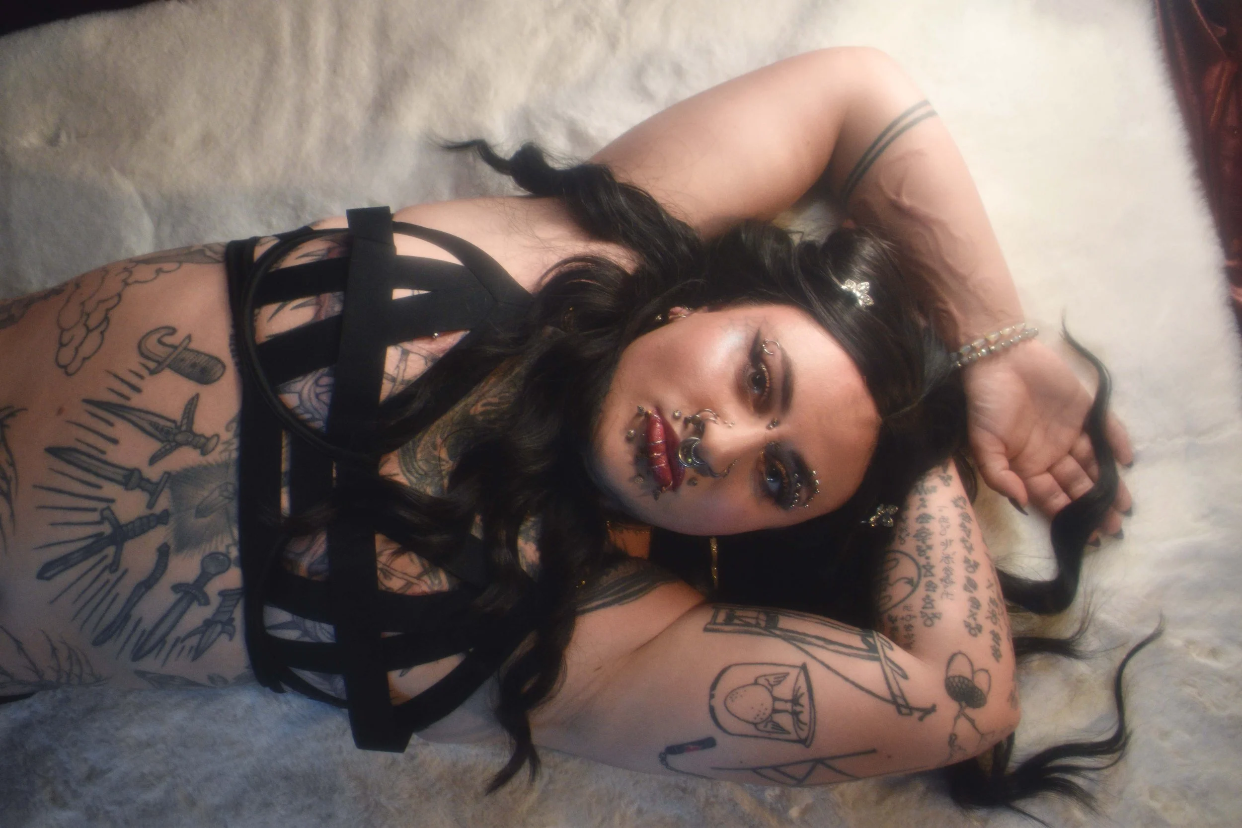 An elite trans luxury escort  lying on her side on a carpeted floor, with long dark hair, tattoos on her arms and chest, and wearing jewelry and black straps. Sexy transgender escort seattle WA Transexual escort for hire San Francisco San Jose CA