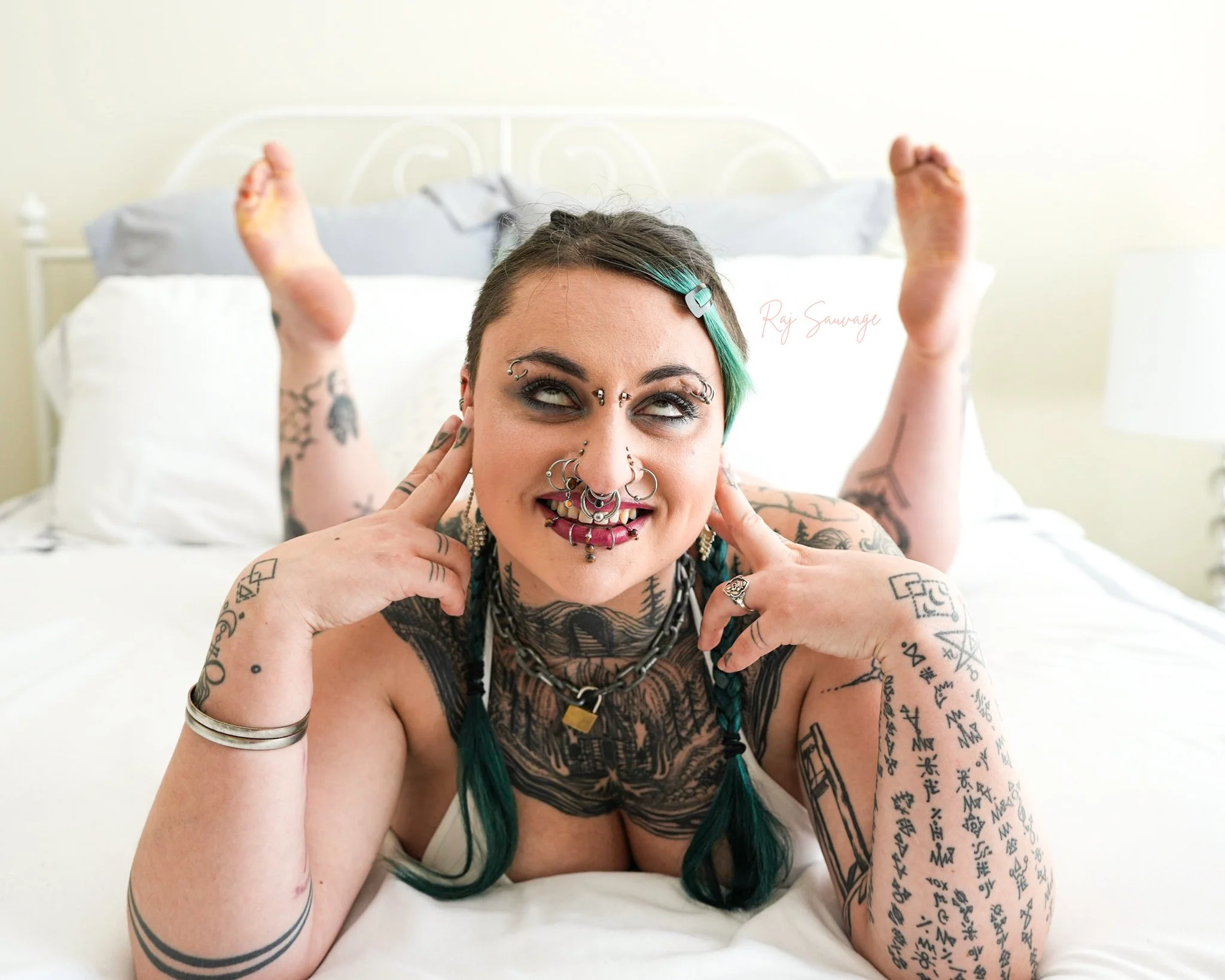 A woman with tattoos and multiple facial piercings lying on a bed, smiling and touching her face, with her legs raised and crossed at the ankles.
