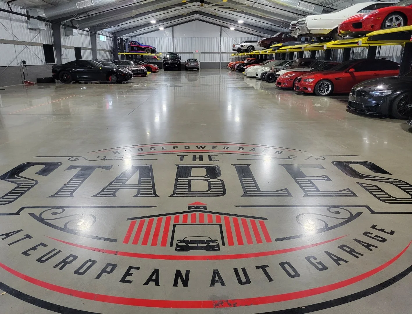 Indoor European auto garage with car storage on elevated racks and a polished concrete floor featuring the logo of The Stables, a horsepower performance brand.