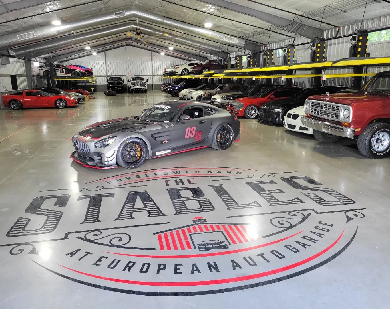 Indoor car storage with various luxury and sports cars, including a matte gray Mercedes-AMG GT Black Series in the foreground, a red Chevrolet Corvette, and a red classic truck on the right. The floor features a large logo reading 'The Stables at European Auto Garage'.