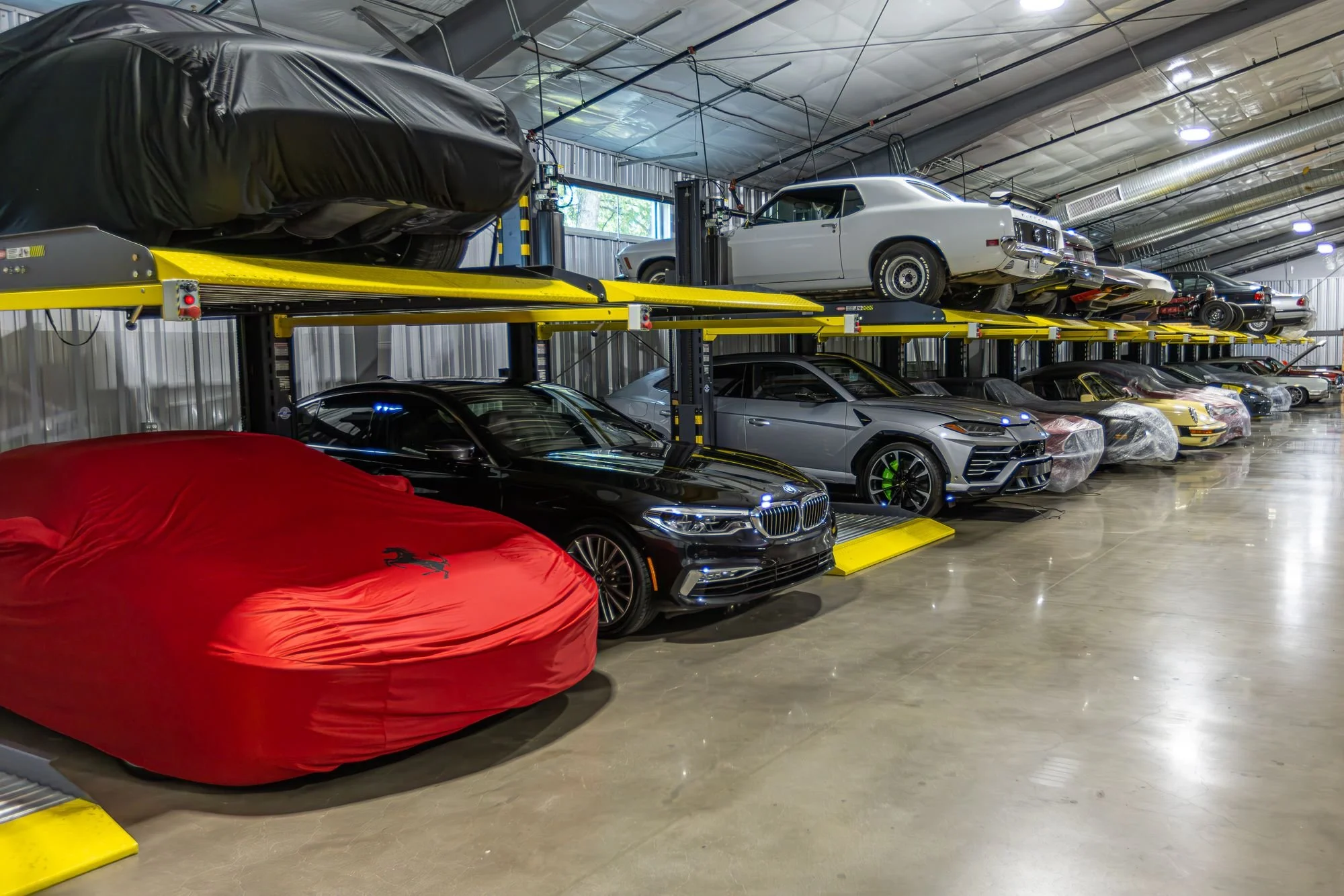 A garage with two levels of luxury cars, some covered and some uncovered, including a black car with a red cover, silver, yellow, and white vehicles, and a partially covered BMW on the lower level.