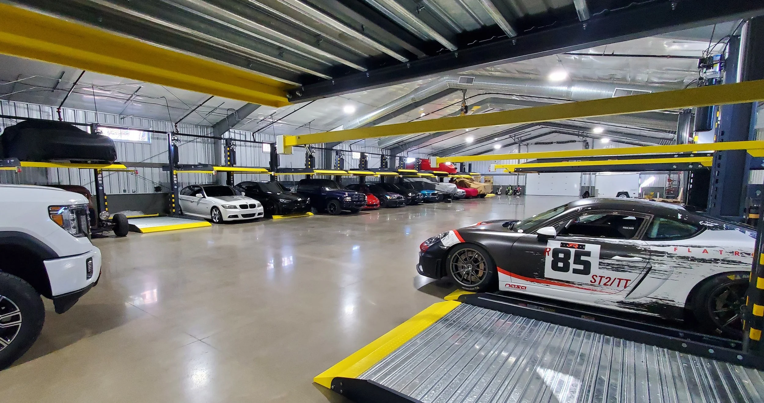 An indoor parking garage with a variety of parked cars, including sports cars, SUVs, and sedans, along with a car lift system and concrete flooring.