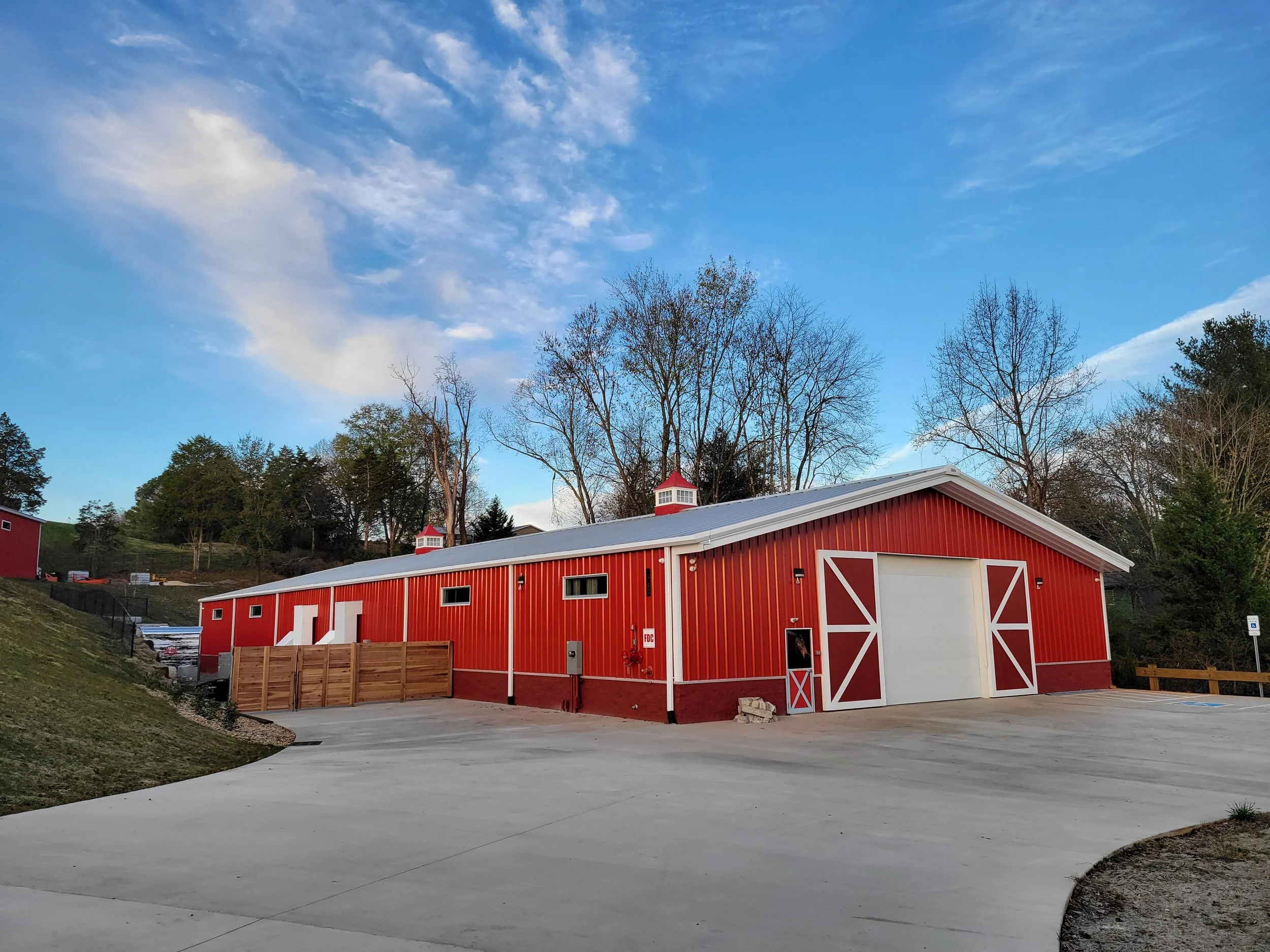 Large red barn with white trim in a rural area, surrounded by trees and a clear blue sky with scattered clouds.