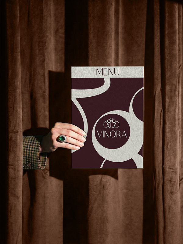 A hand with a large green ring holding up a menu with the word "VINORA" and a logo, peeking through brown curtains.