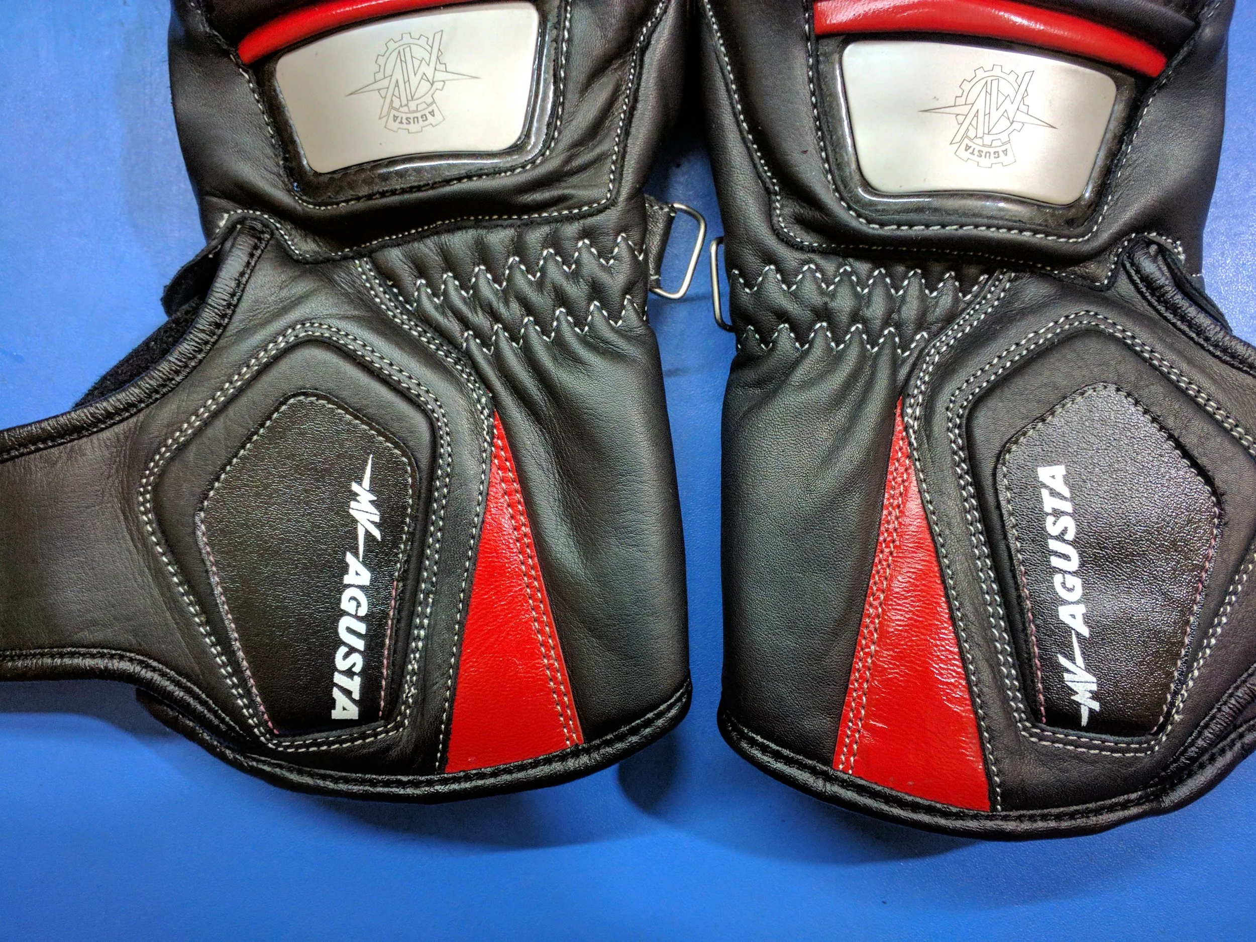 Close-up of a pair of black and red racing gloves with white stitching, displaying the logo 'AUSTIN 4G' on the black leather surface. The gloves are laid flat on a blue background.