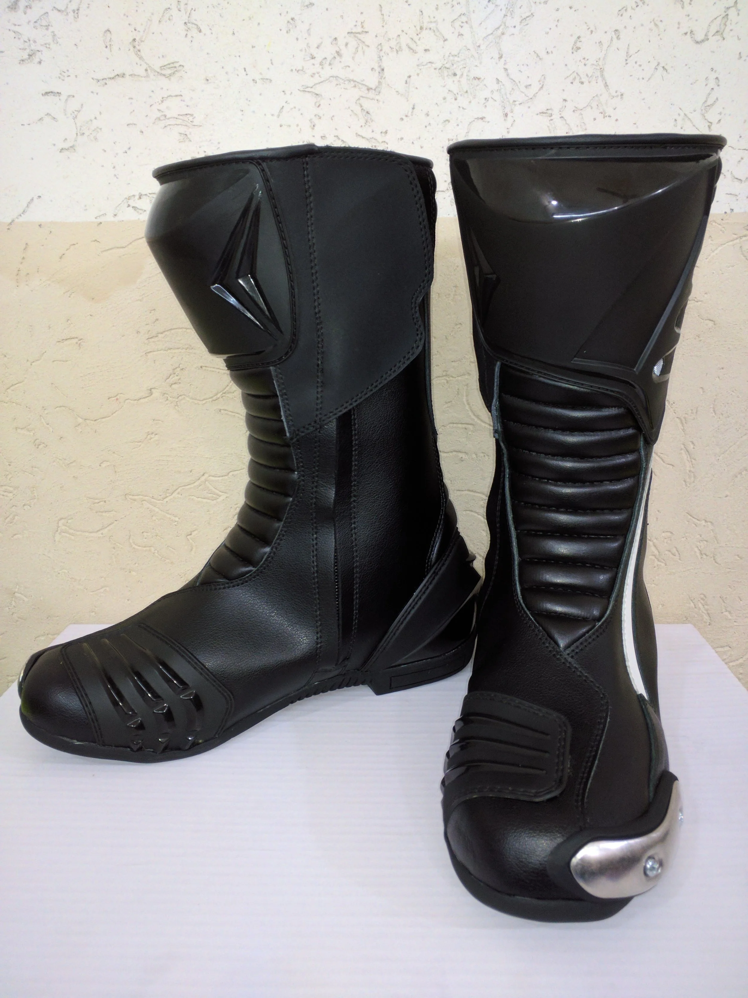 Pair of black motorcycle boots with shiny tips and stitched leather design, placed on a white surface against a beige textured wall.