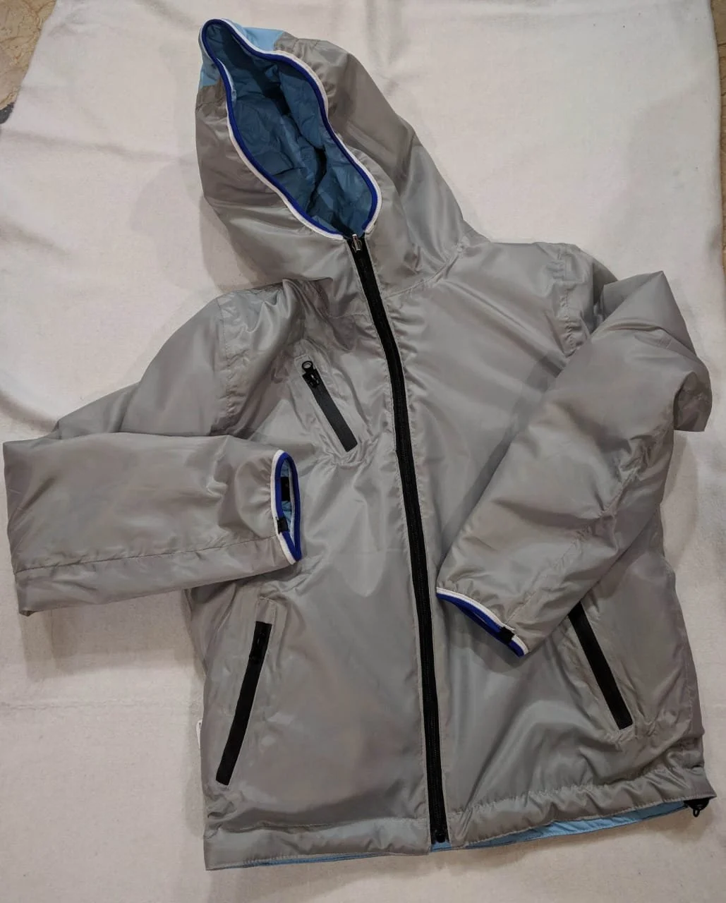 Gray outdoor jacket with zipped pockets and a blue-lined hood, laid on a white fabric surface.