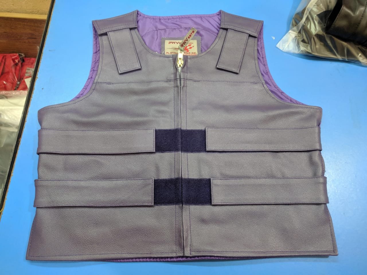A gray tactical vest with three horizontal straps on a blue surface, with purple interior lining and a front zipper.