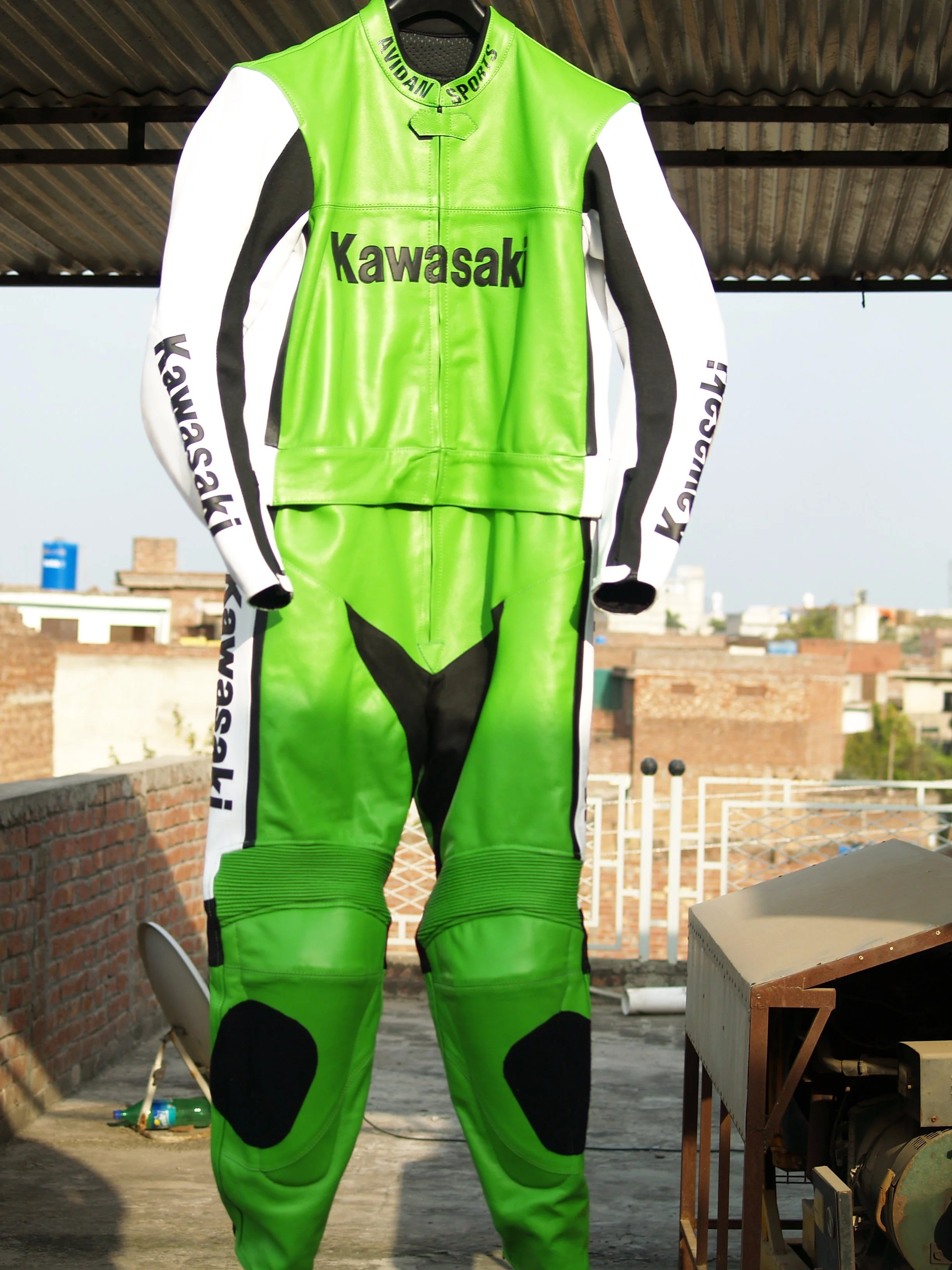 Green and white Kawasaki sports motorcycle racing suit hanging in a rustic outdoor space with brick walls and a corrugated metal roof.