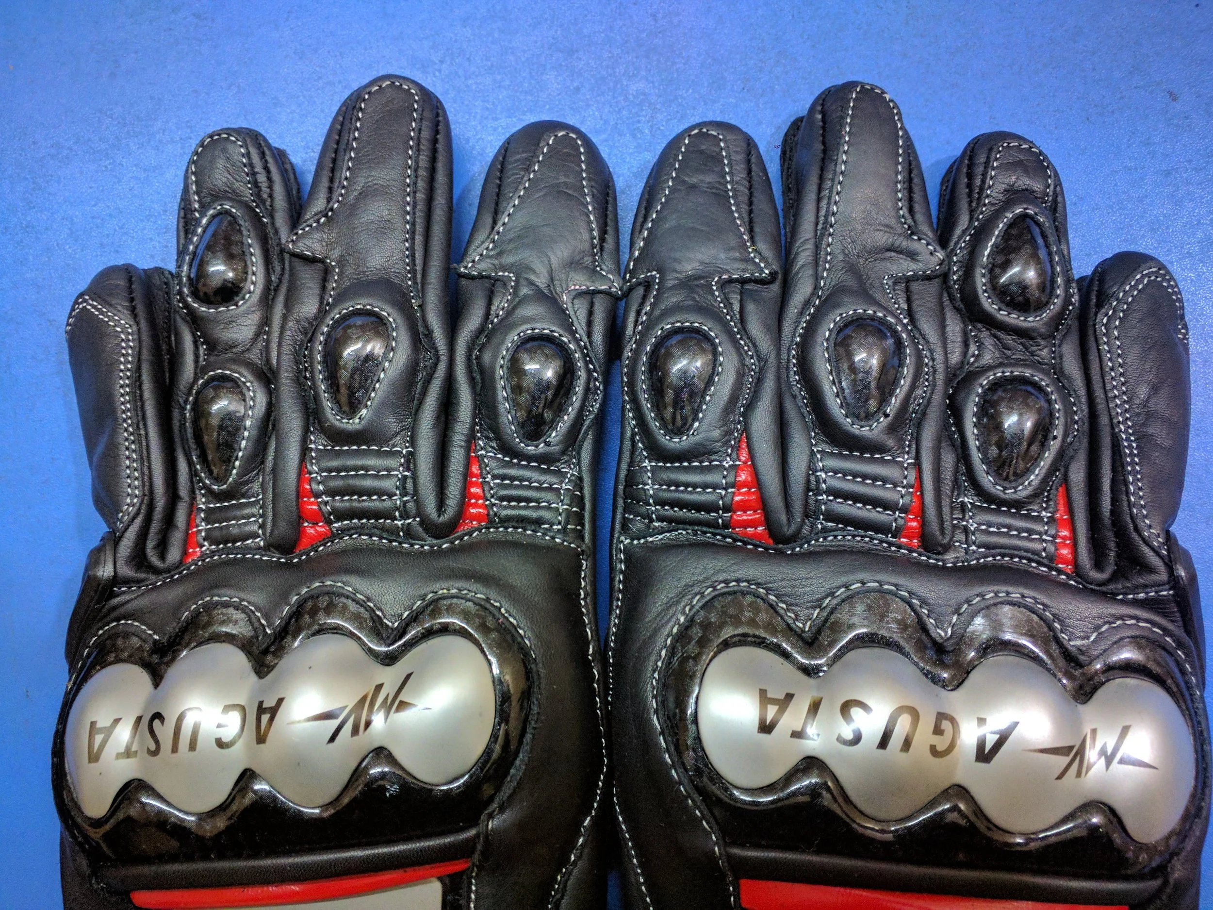 Close-up photo of black leather driving gloves with white stitching and some metallic detailing on the fingers, placed on a blue surface.