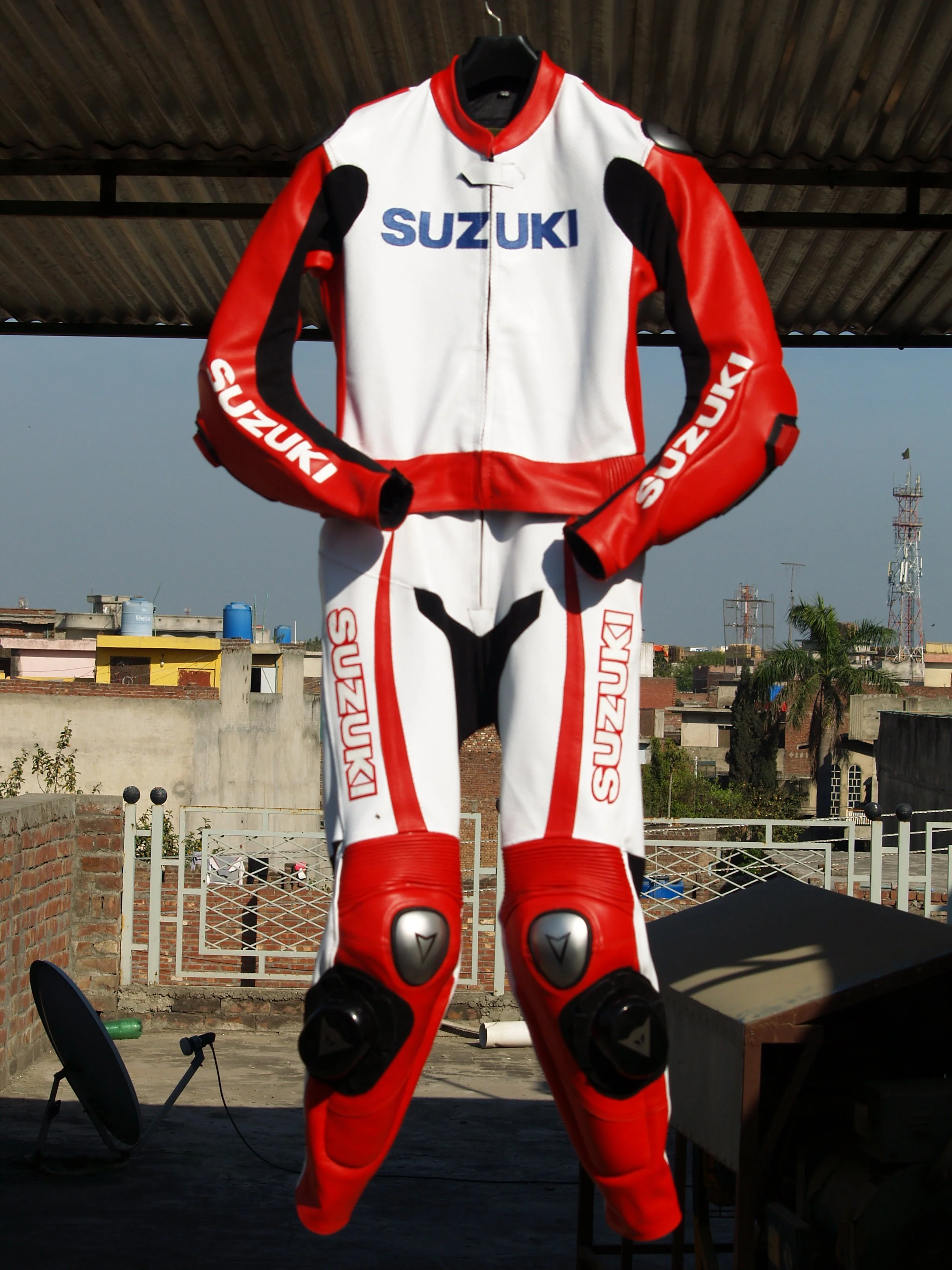 A red and white Suzuki motorcycle racing jacket with black accents, hanging on a hanger. The background includes a rooftop view of a cityscape with buildings, rooftops, and communication towers.