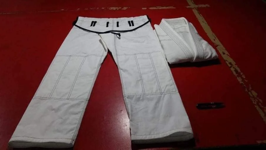 A pair of white martial arts Gi pants with black drawstring, laid flat on a red mat, next to folded white Gi top and a black marker.