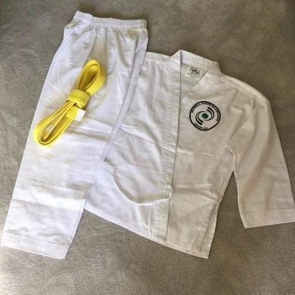 A white martial arts gi with a yellow belt placed on top of the pants, laid out on a gray carpeted surface.