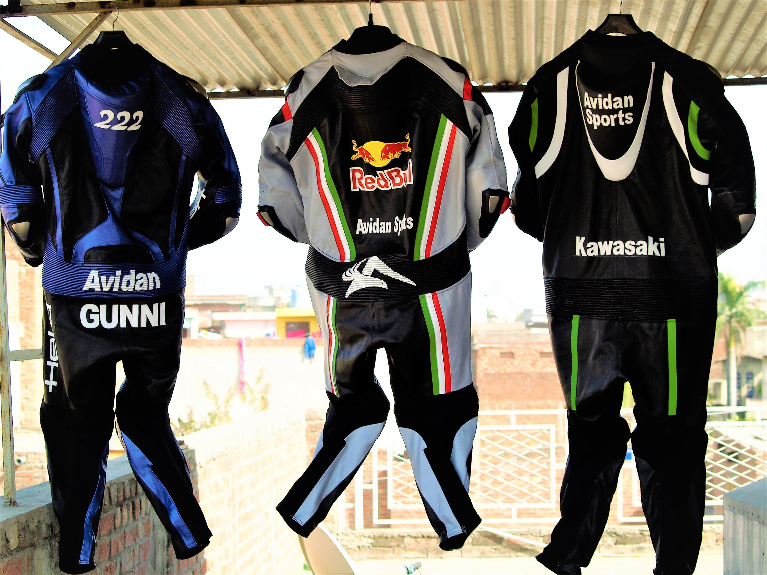 Three motorcycle racing suits hanging on hangers from a rod in a balcony or veranda with a metal roof overhead, with buildings and sky in the background.