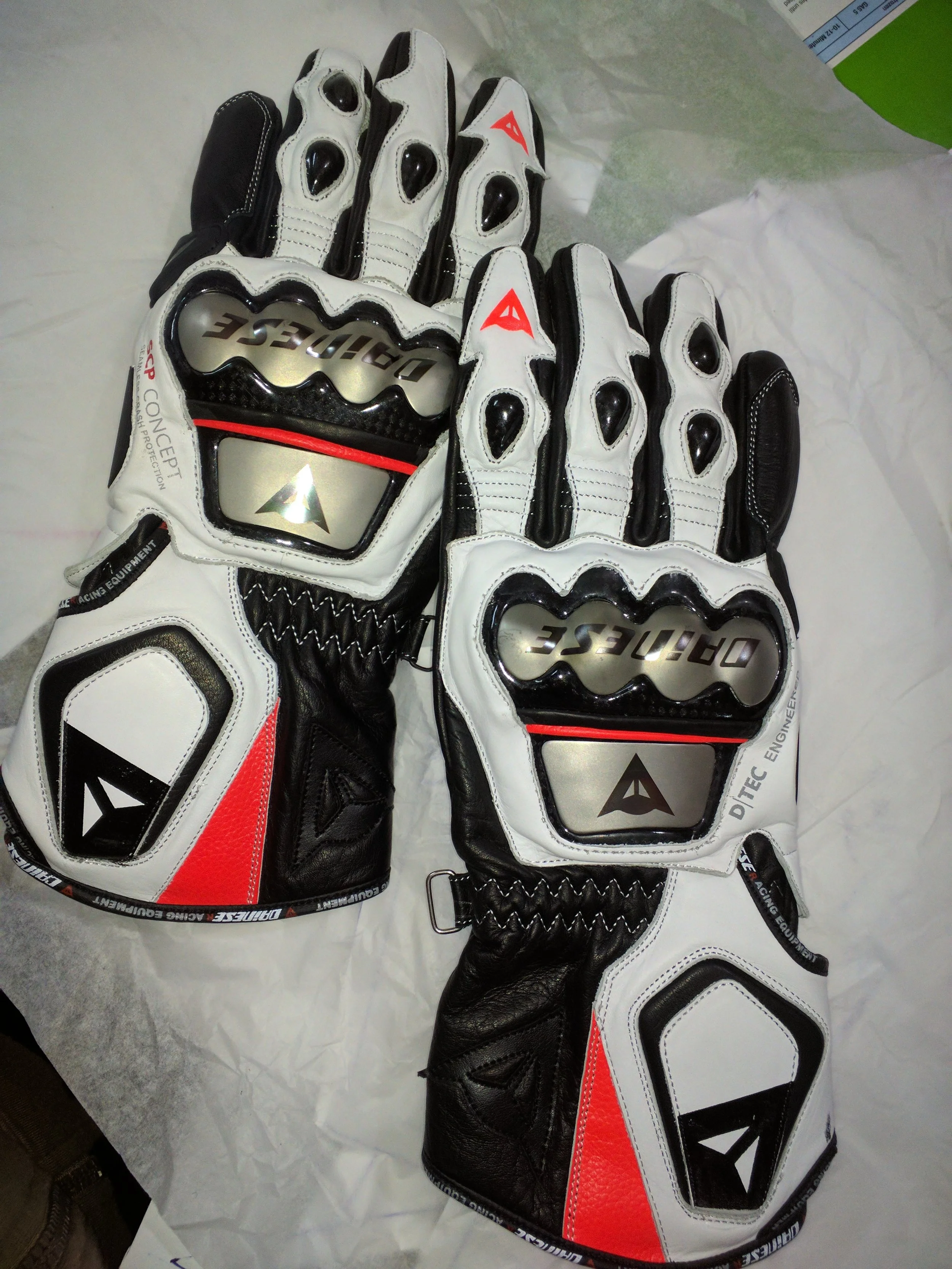 Pair of racing gloves with black, white, red, and gray sections, featuring protective knuckle pads and branded with Ditech and Alpinestars logos, laid on tissue paper.