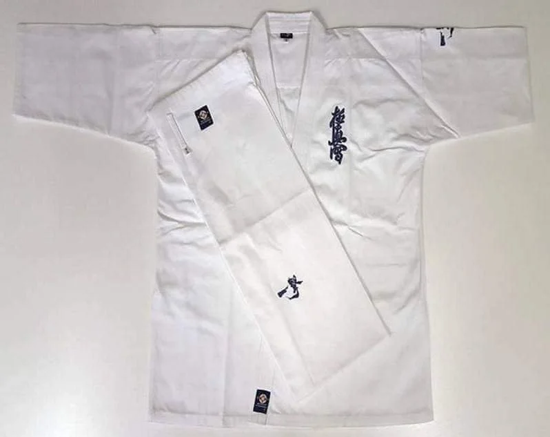 A white martial arts uniform, called a gi, laid flat on a surface. The gi has blue embroidery on the chest and an embroidered patch on the waist area, with a small black and white tag on the collar and a patch on the sleeve.