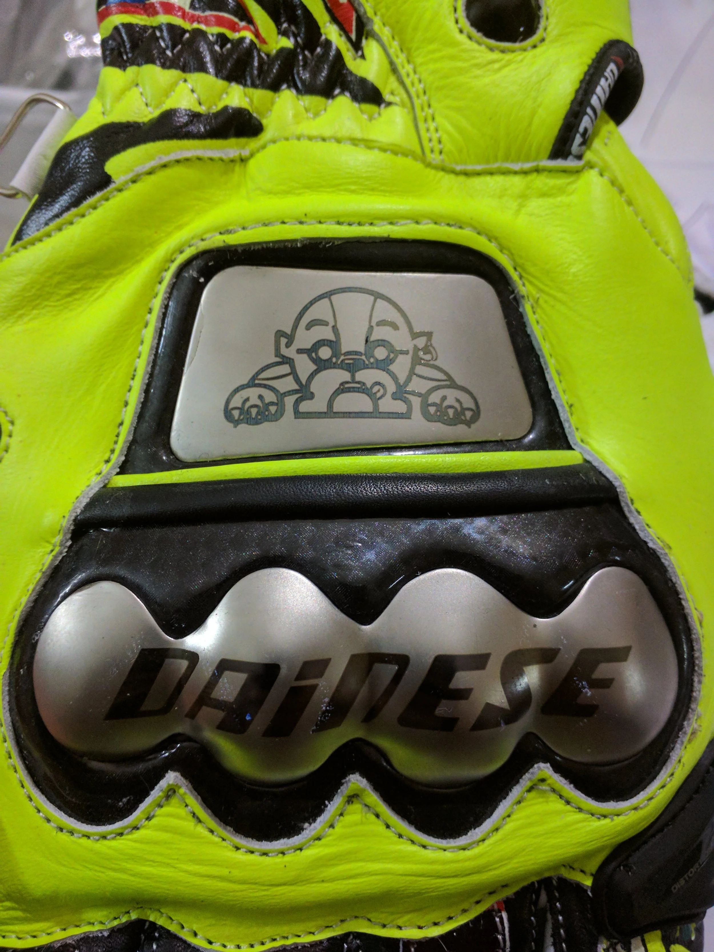 Close-up of a yellow and black sports glove with a metallic plate depicting a cartoon baby skull with a pacifier, and the word "DARE" partially visible.
