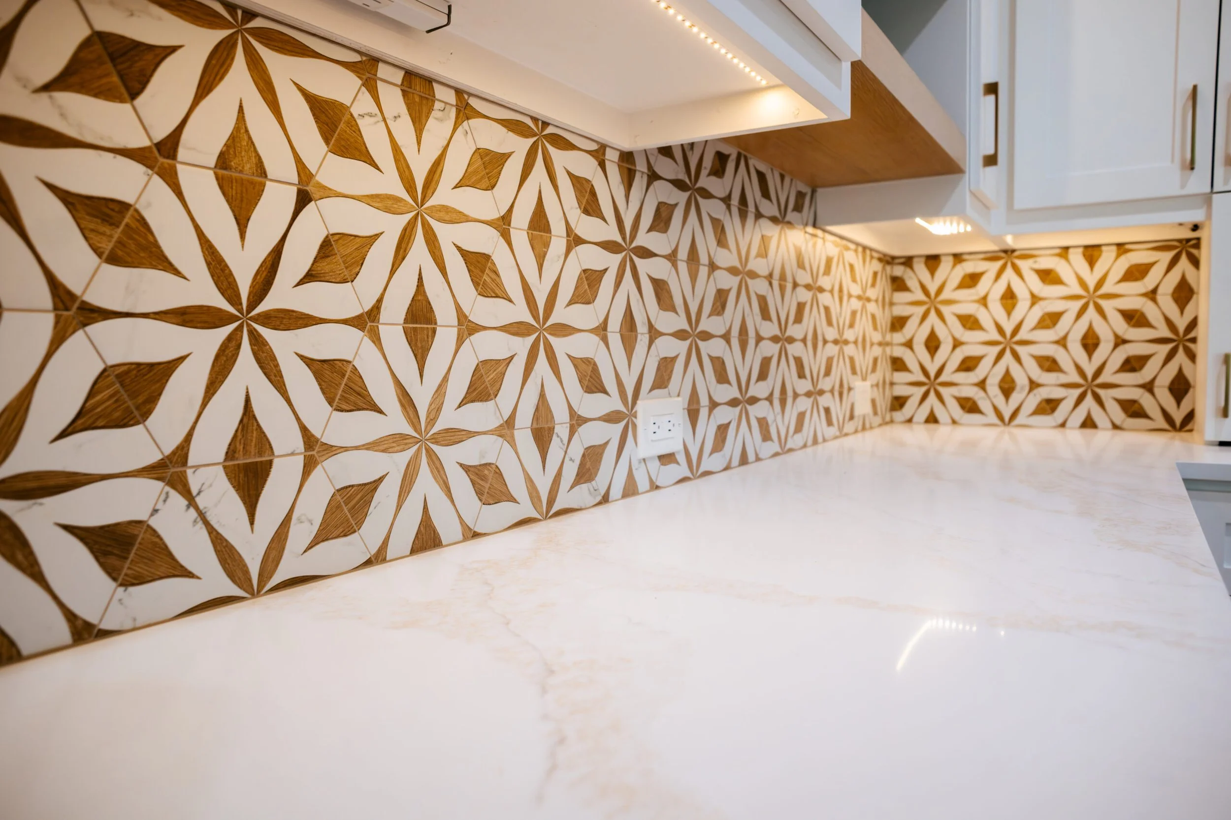 Close-up of a decorative kitchen backsplash with a geometric pattern of brown and white tiles, a white countertop with a power outlet, and part of cabinets and lighting fixtures.