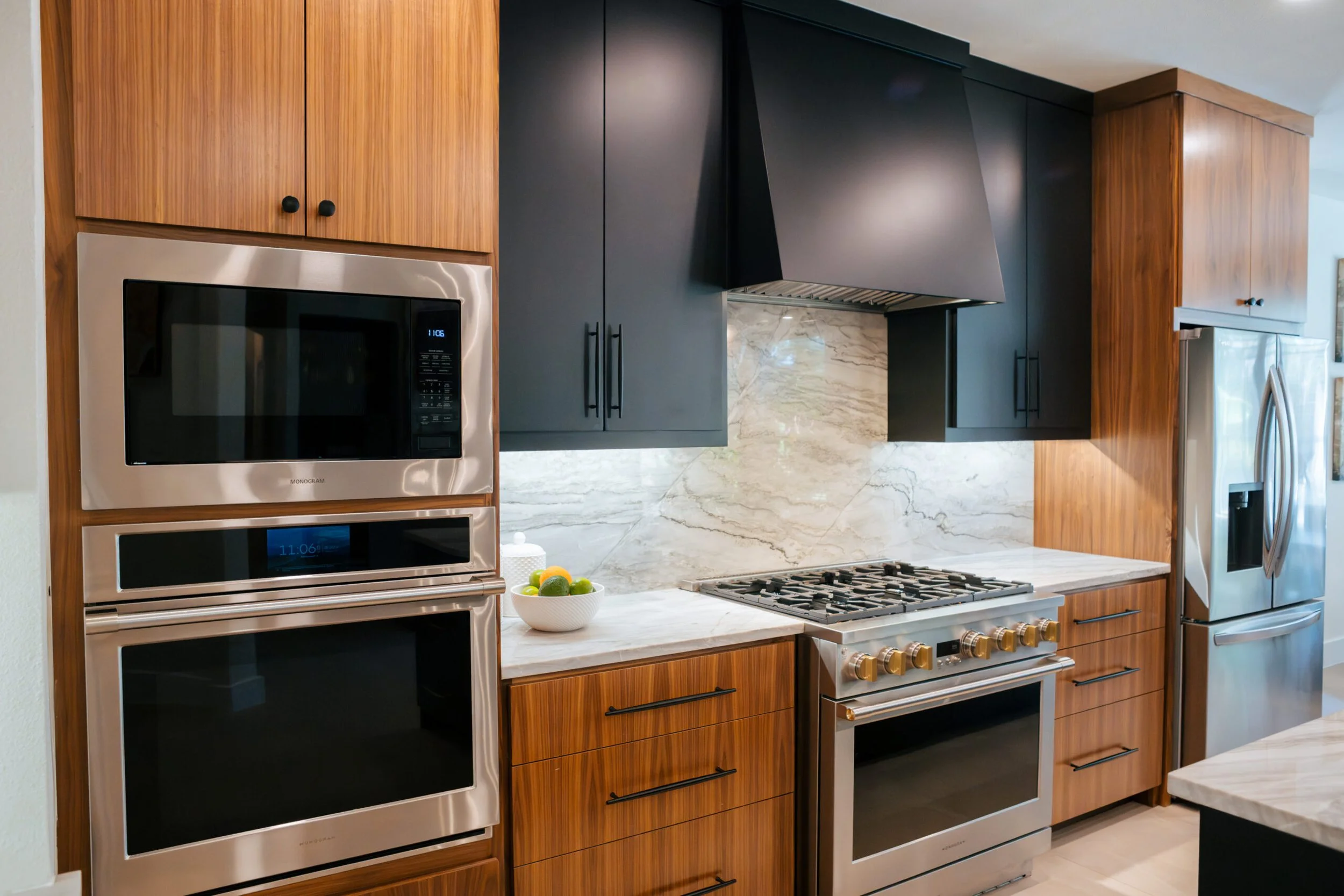 Modern kitchen with black and wood cabinetry, stainless steel appliances, marble backsplash, and a bowl of lemons and limes on the countertop.