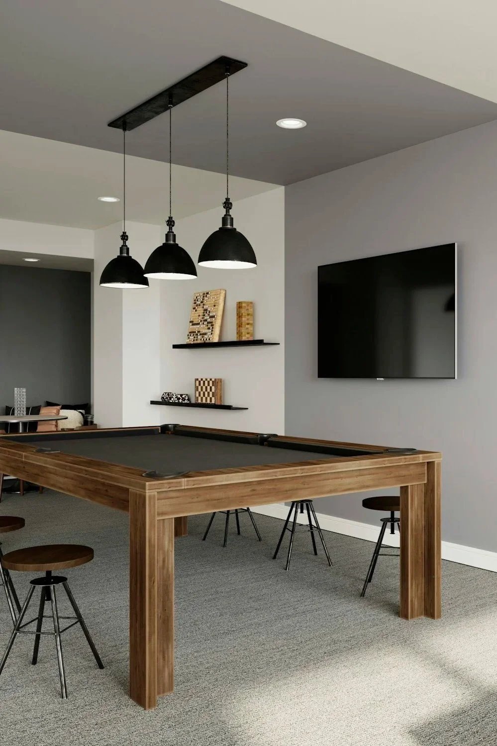 Interior of a modern game or recreational room featuring a wooden pool table, black pendant lights above, a mounted television on a light gray wall, and shelves with decorative items.