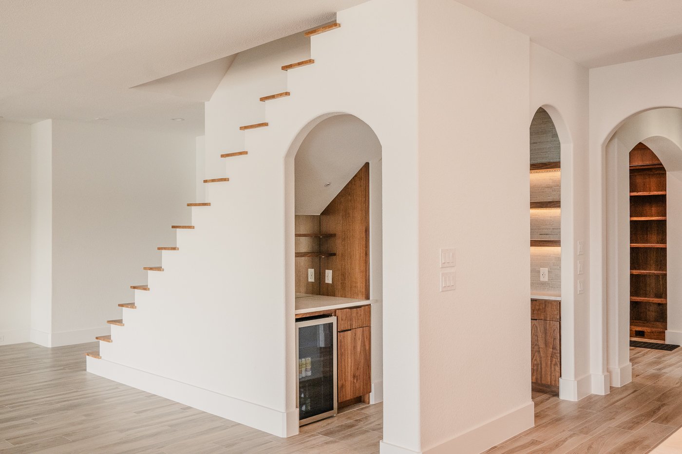 Interior view of a modern home with white walls, wooden flooring, and a staircase with open wooden steps. The space includes a small built-in bar with shelves and cabinets, and arched doorways leading to additional rooms with wooden features.