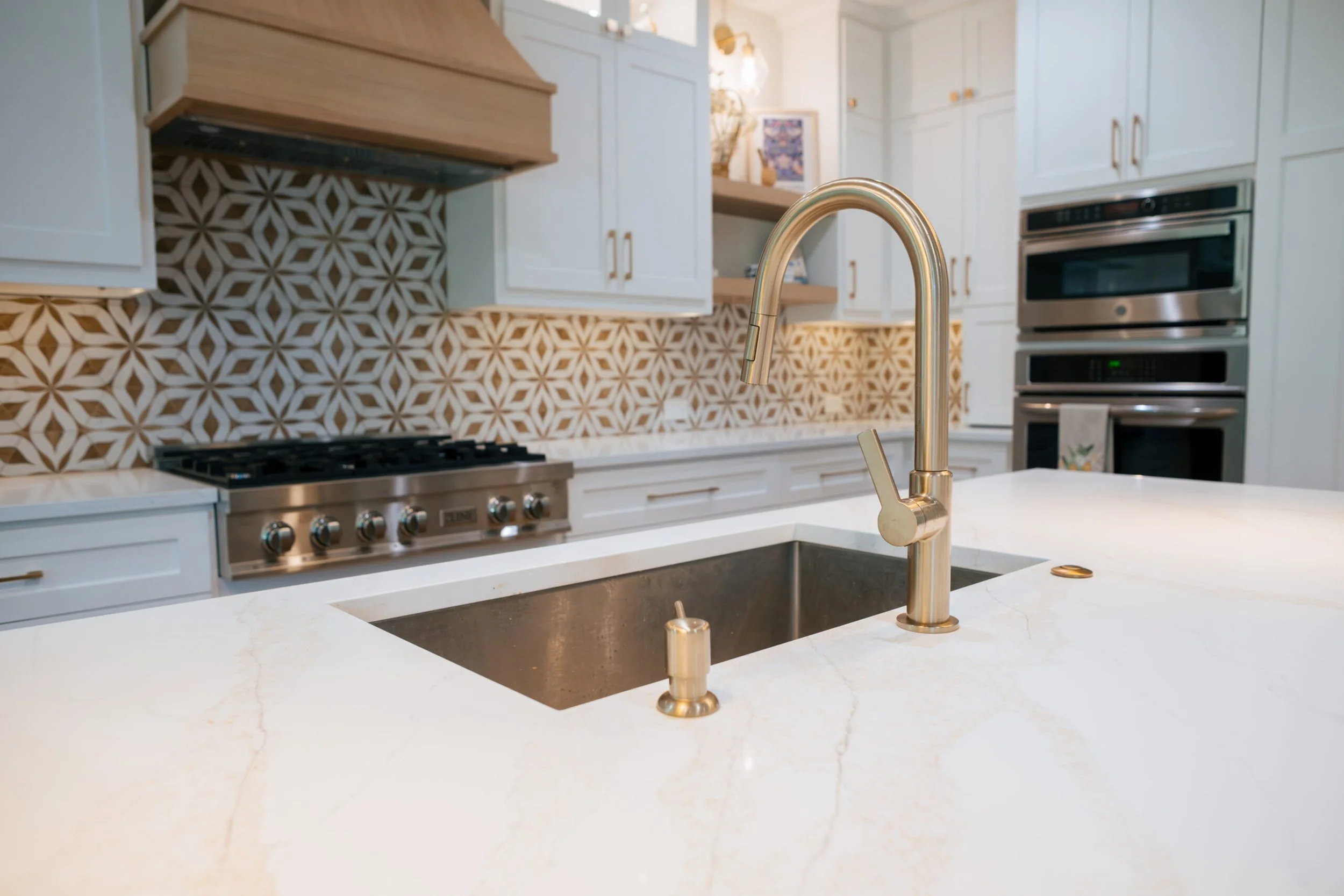 Close-up of a modern kitchen sink with a gold-colored faucet on a white marble countertop, with patterned tile backsplash and white cabinets in the background.