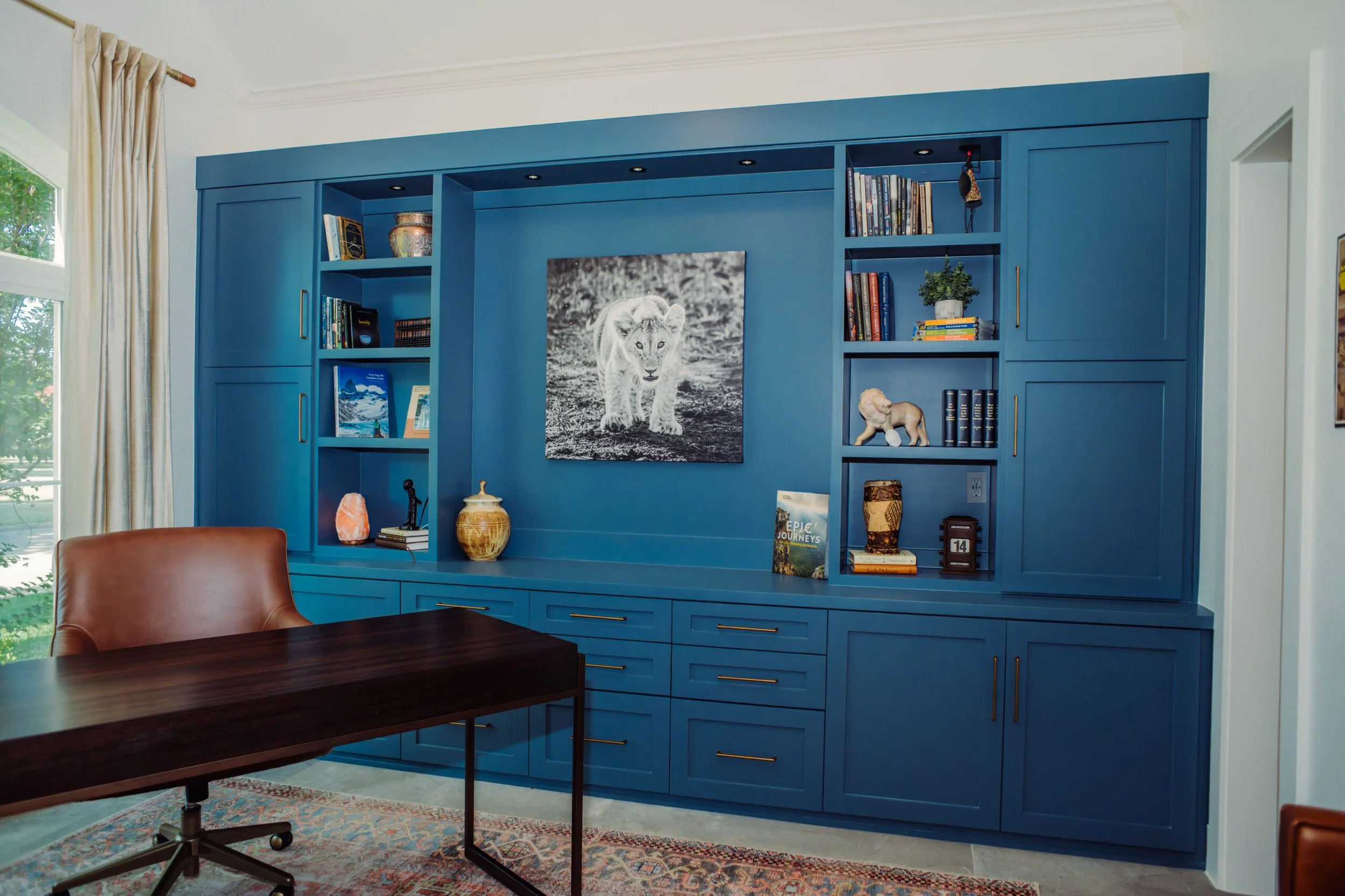 A blue built-in bookshelf with books, decorative items, and a framed photo of a lion cub in a living room with large window and wooden desk.