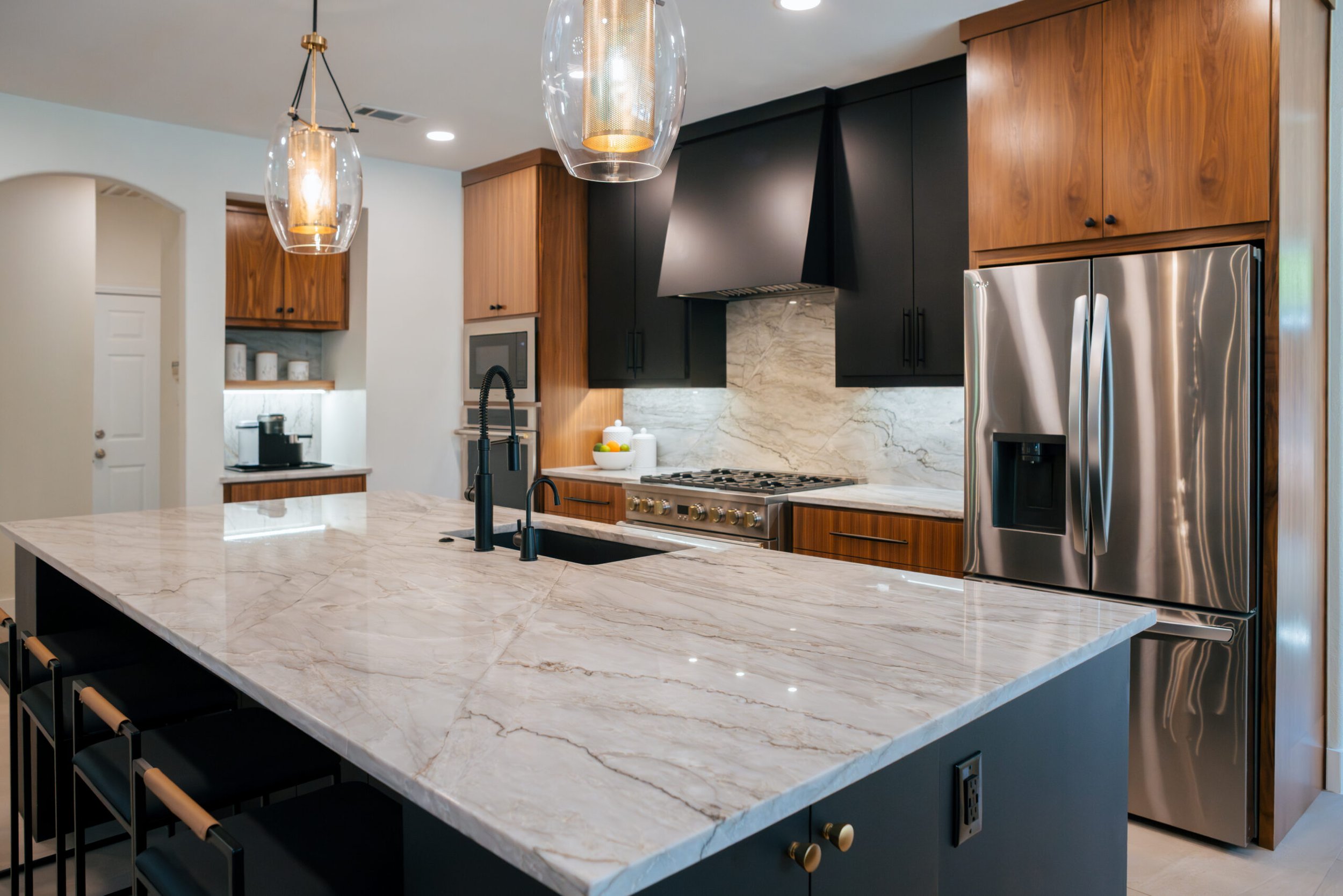 Modern kitchen featuring a large marble island with a black sink and gold faucet, built-in oven, stainless steel refrigerator, and wooden cabinets with black accents, illuminated by pendant lights.