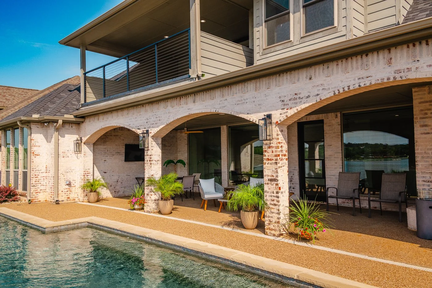Exterior view of a house with a patio by the pool, featuring white brick walls, arched openings, potted plants, outdoor chairs, and a balcony above, with a lake in the background.