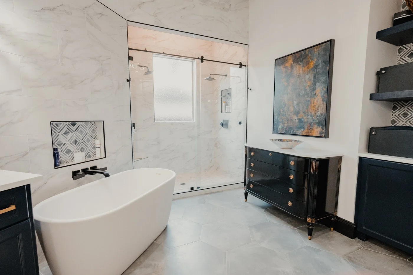 Modern bathroom with a freestanding bathtub, glass shower enclosure, black dresser, abstract artwork, and a window with frosted glass.