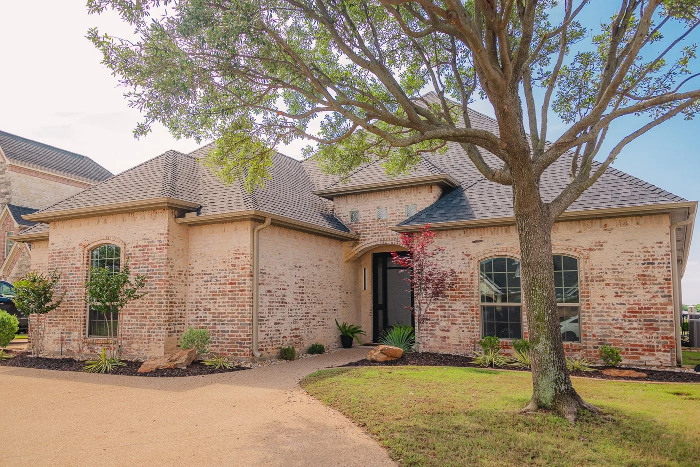 Brick house with a large tree in the front yard, two windows on the right and a small landscaped garden with rocks and plants in the front.